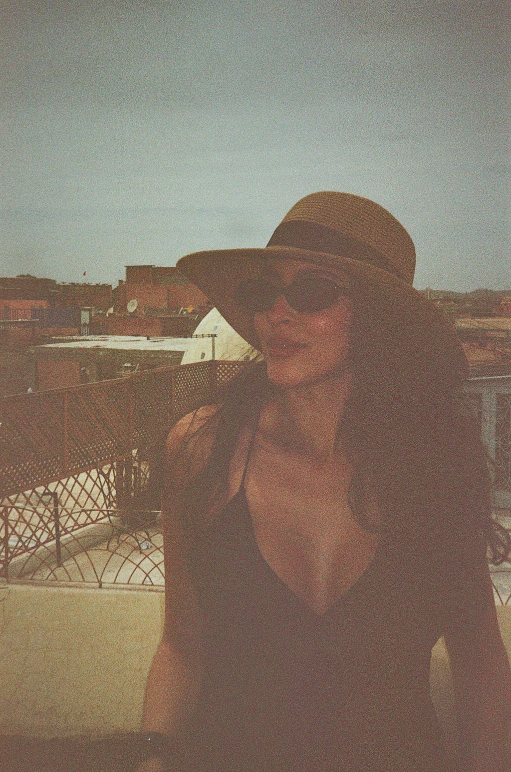 A stylish woman wearing a wide-brim hat and sunglasses poses on a rooftop terrace in warm sunlight.