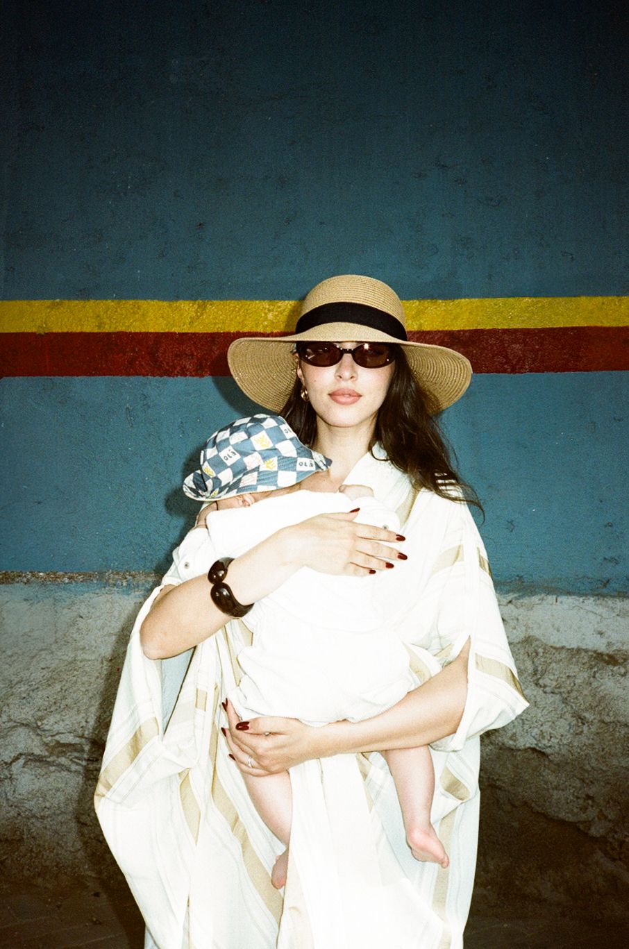 Woman in straw hat and sunglasses holding a baby in white wrap against colorful blue wall.