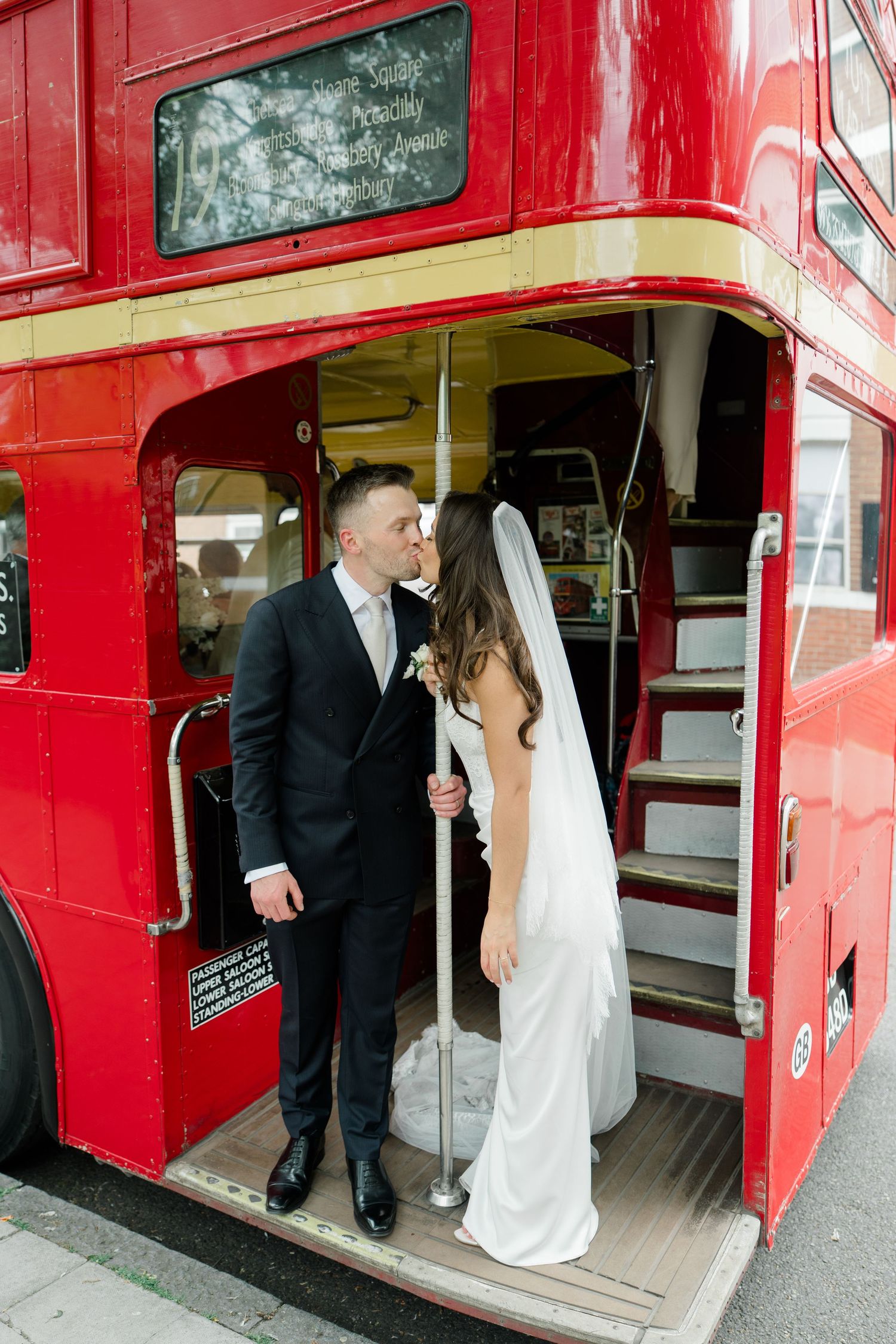 Bride and groom share a kiss on the platform of a classic red London double-decker bus on their wedding day.