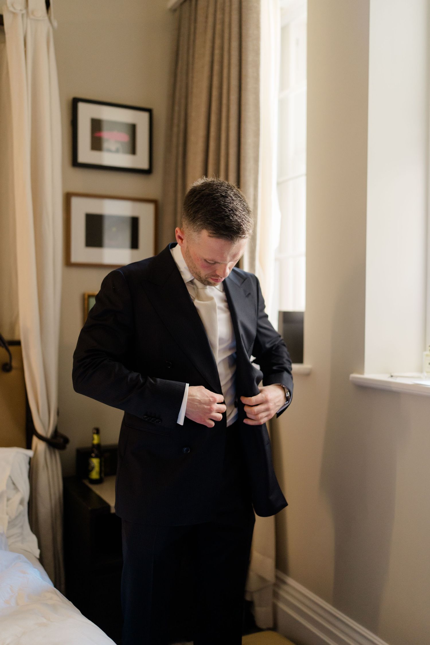 Groom buttoning his dark suit jacket while getting ready near a window in an elegant hotel room.