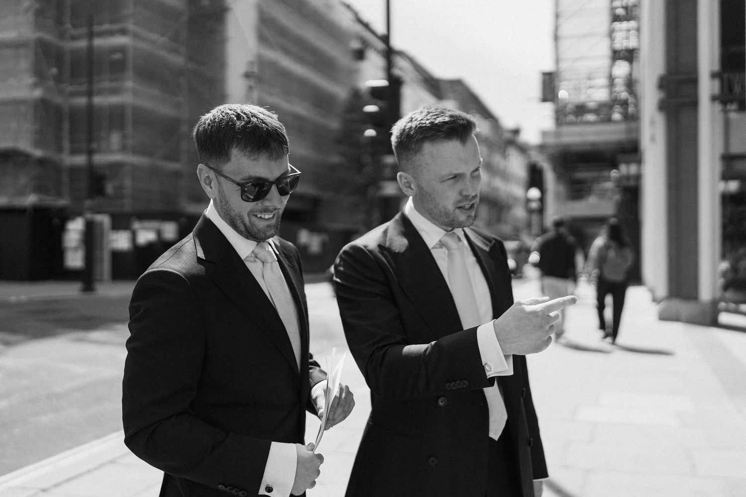 Two groomsmen in black suits walking outdoors on wedding day in black and white.