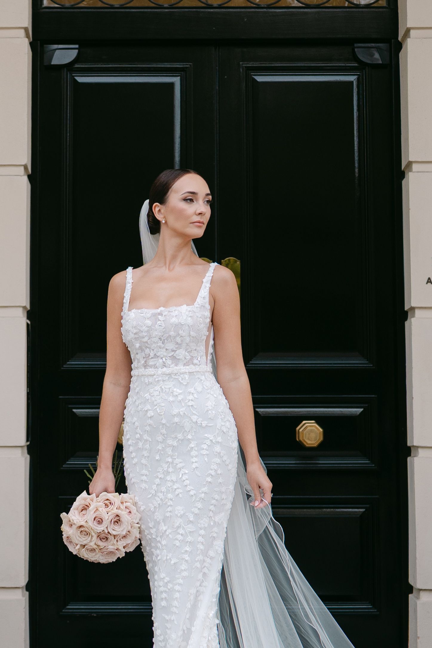 Stunning bride in fitted lace dress poses against black door with blush bouquet.