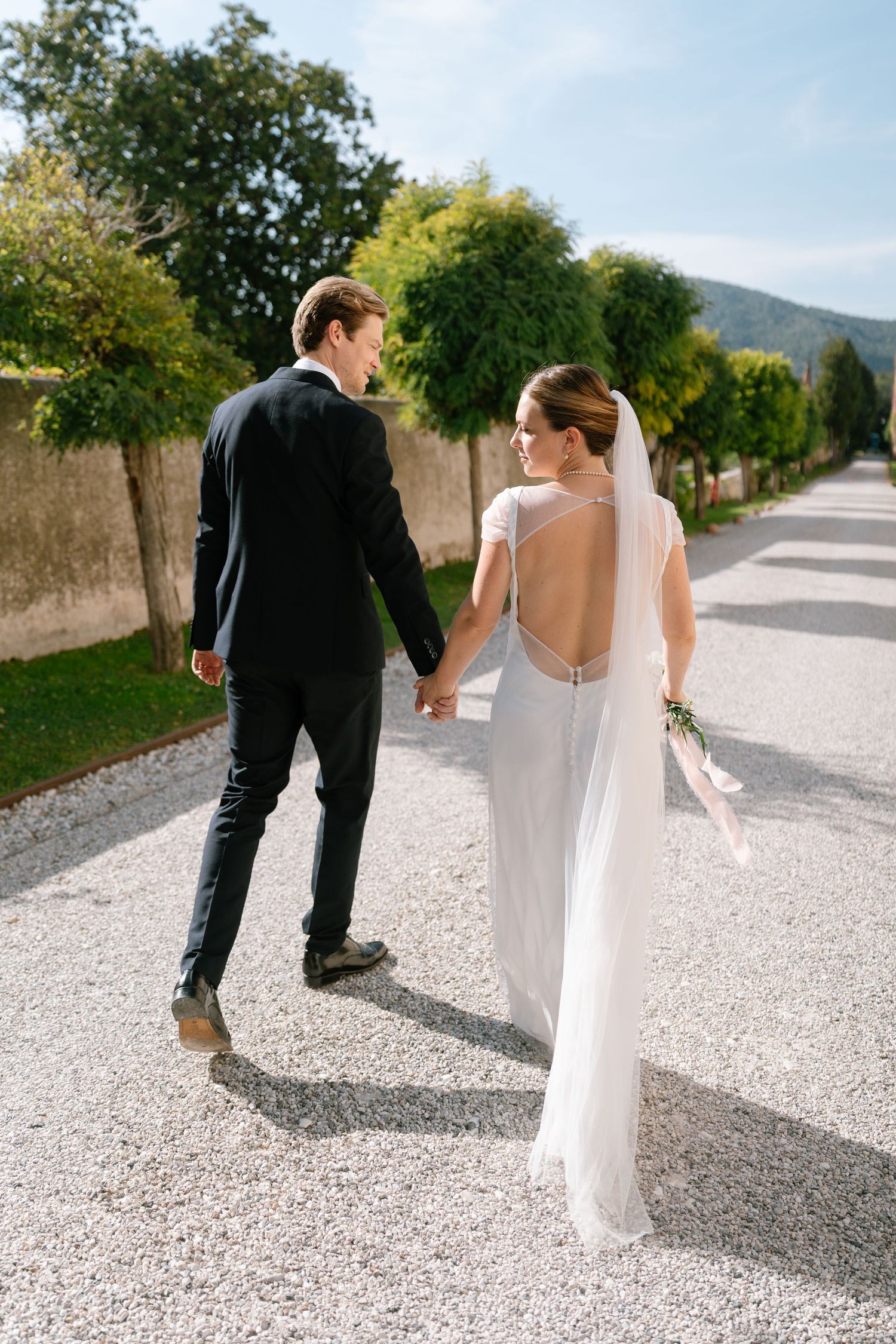 Couple walking hand in hand on path, bride wearing elegant backless wedding dress.