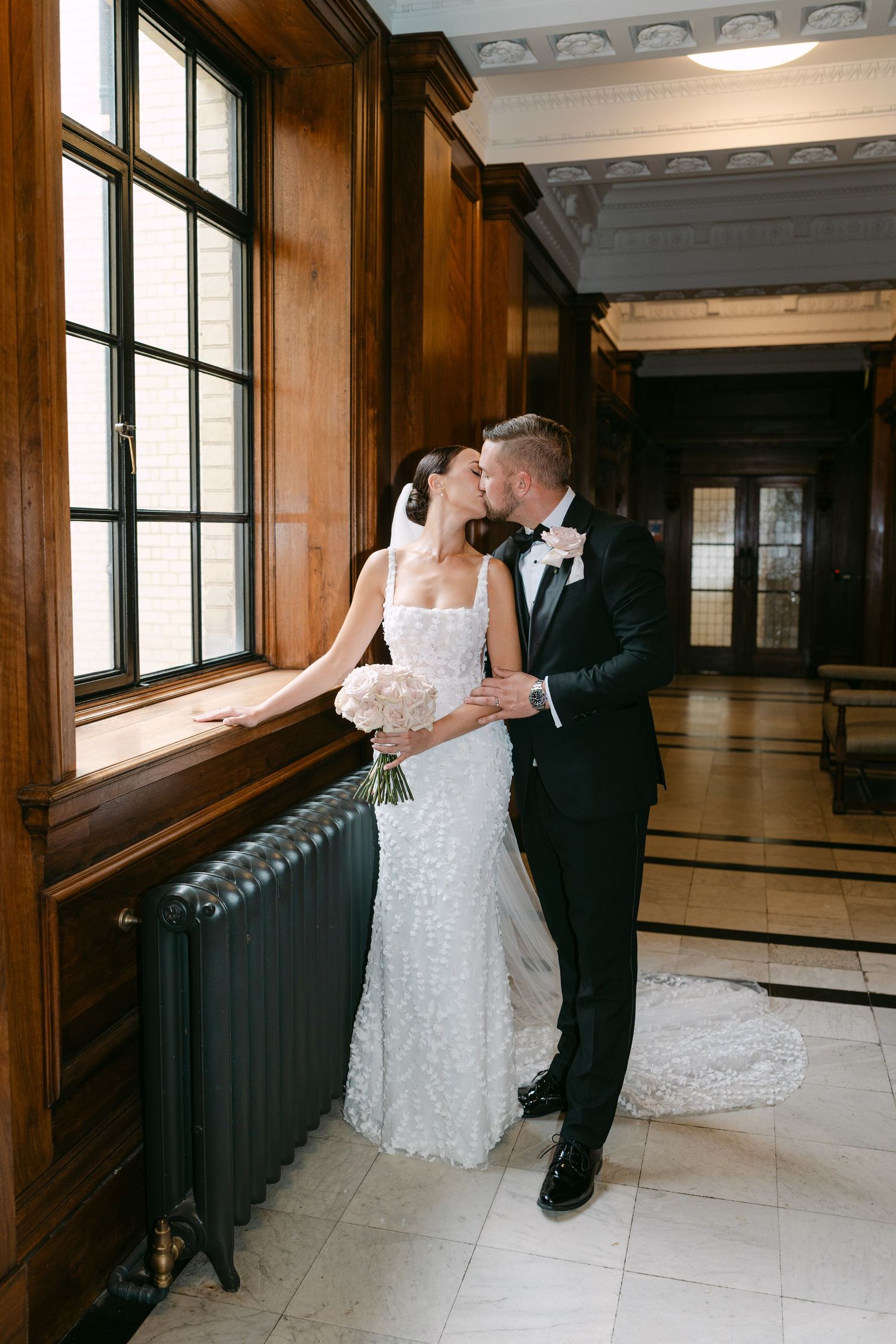 Wedding couple embraces by large windows in an ornate hallway with vintage radiators.