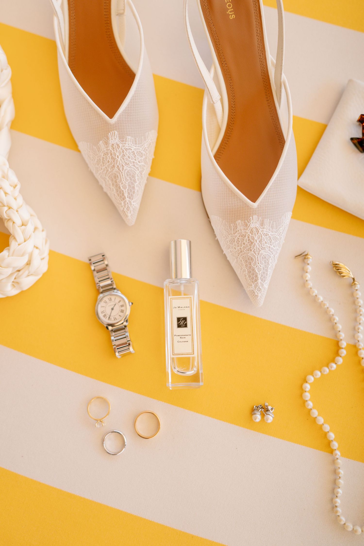 Wedding accessories including white lace shoes, watch, perfume and jewelry on yellow striped background.