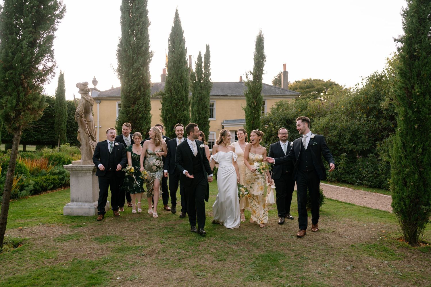Wedding party members gather for group photos on landscaped grounds with cypress trees.