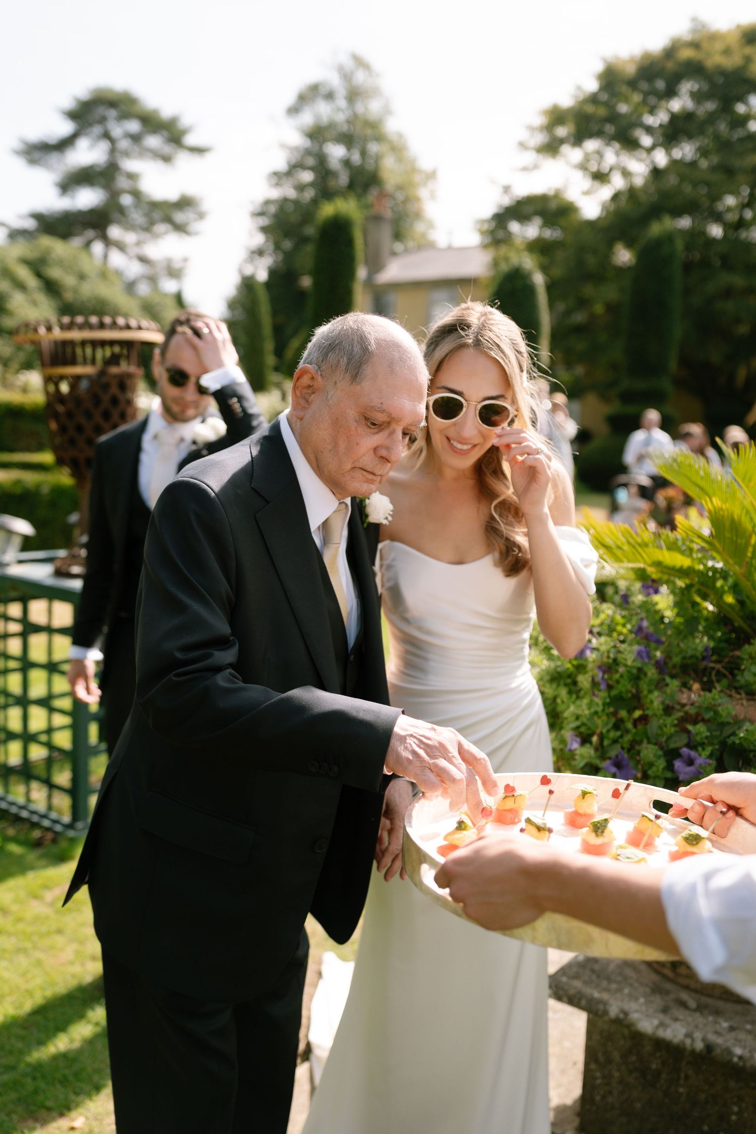 Guests examine appetizers being served on a silver platter in a sunny garden setting.