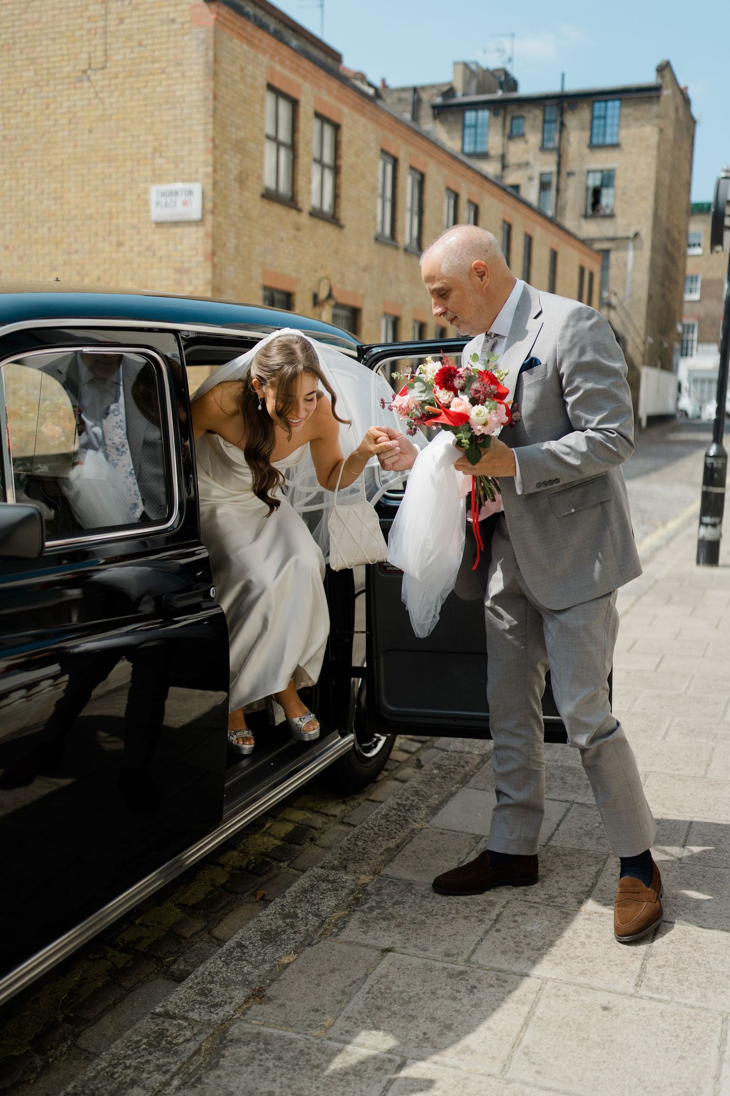Groom in grey suit helps bride in white dress exit a vintage black car on a sunny cobblestone street.