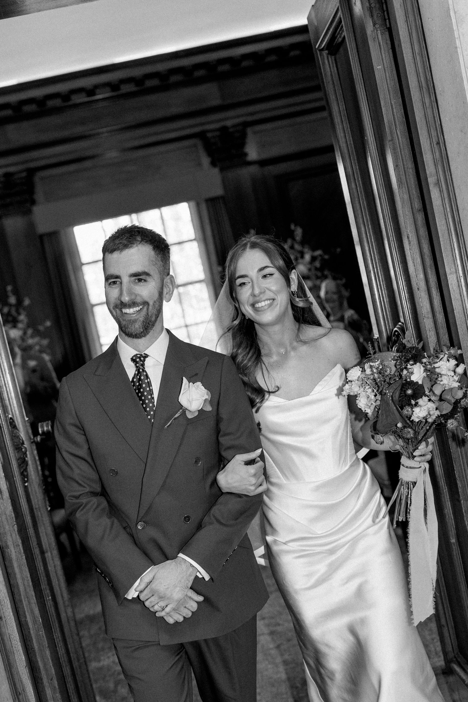 Black and white photo of smiling bride and groom posing in doorway, bride holding bouquet in elegant satin gown.