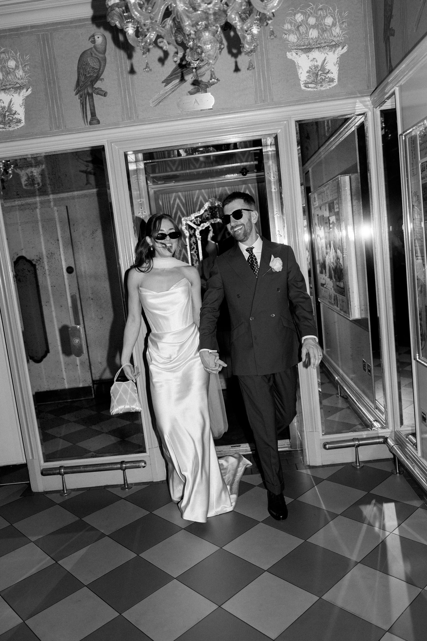 Black and white photo of newlyweds in sunglasses walking through hotel lobby.