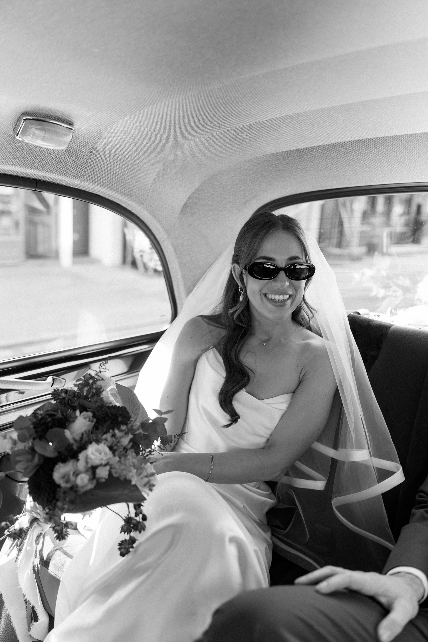 Smiling bride wearing sunglasses and a veil sits in a vintage wedding car holding a bridal bouquet.