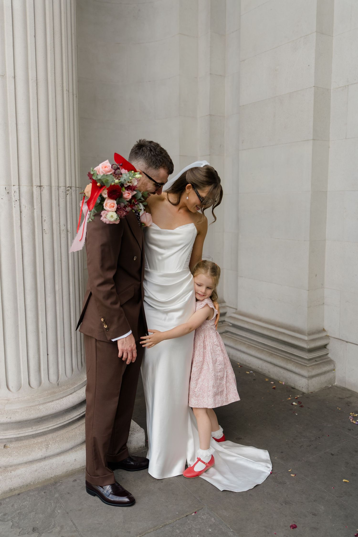 Bride and groom share a kiss while young girl in pink dress hugs bride beside stone column.