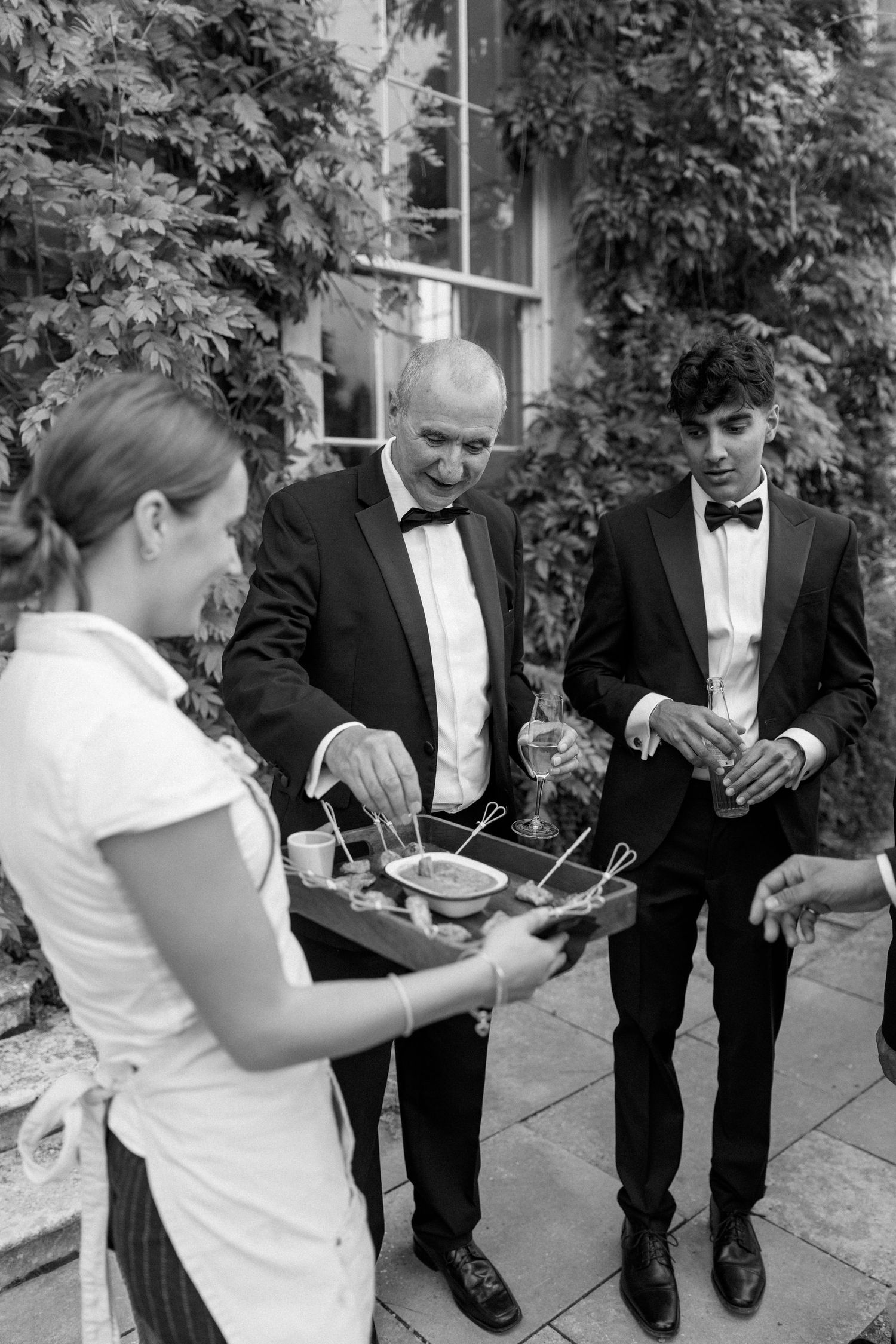 Wedding guests in tuxedos sampling hors d\u0027oeuvres from a tray served by staff at outdoor reception.