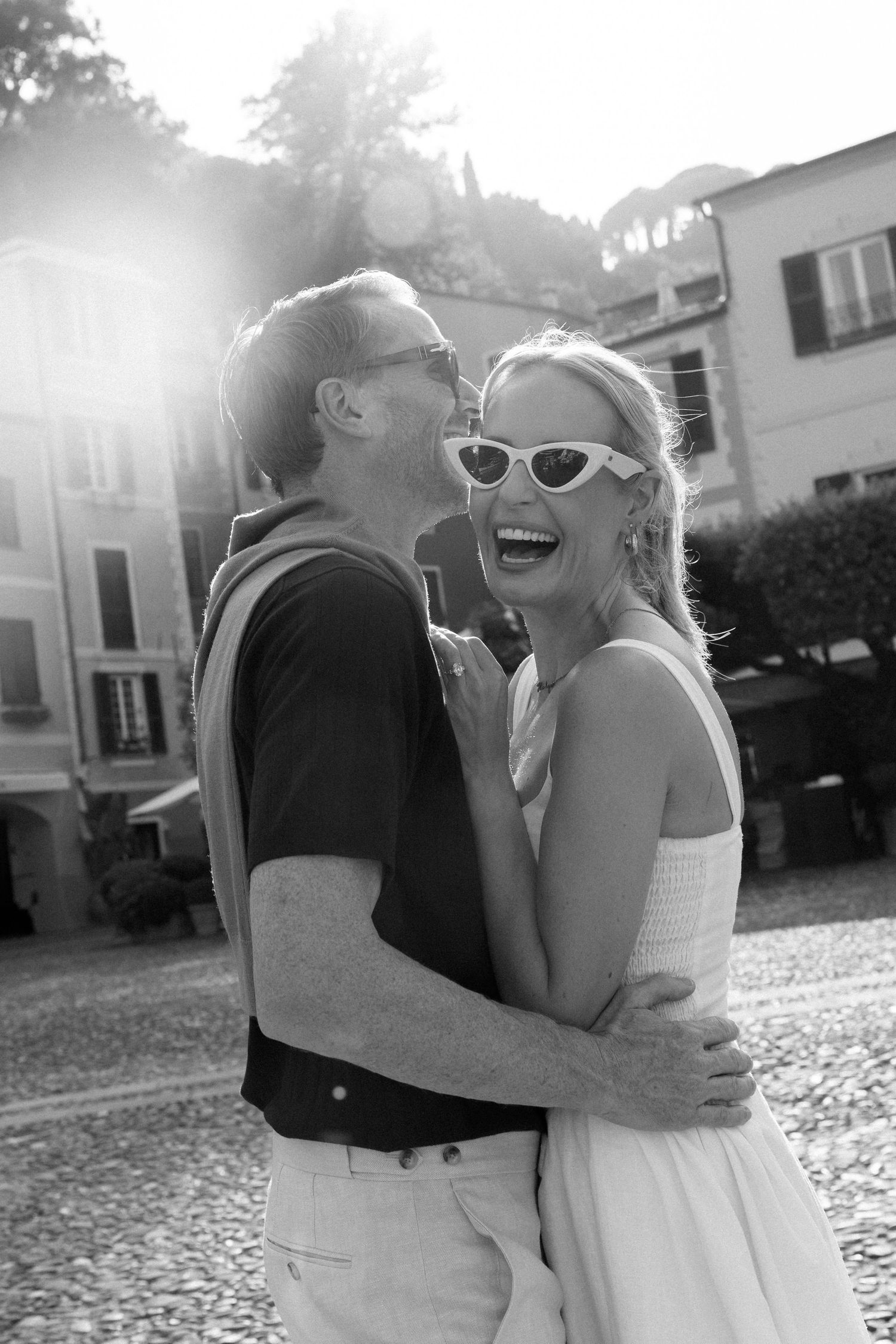 Joyful couple sharing a laugh in bright sunlight on Italian cobblestone plaza, black and white photo.