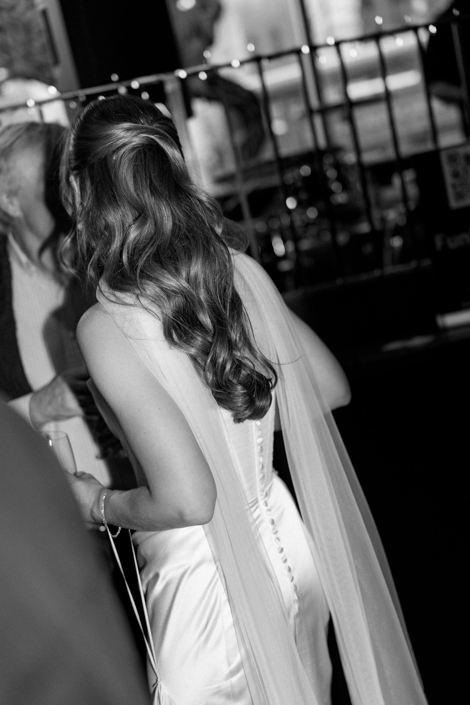 Black and white photo of bride from behind showing long wavy hair, veil, and elegant satin wedding gown.