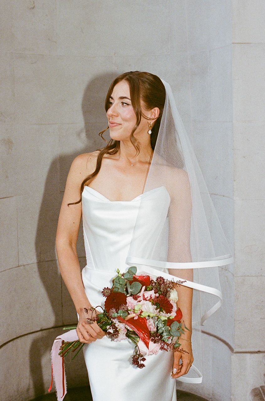 Bride in strapless white gown and veil holding red and green bouquet, smiling softly indoors.