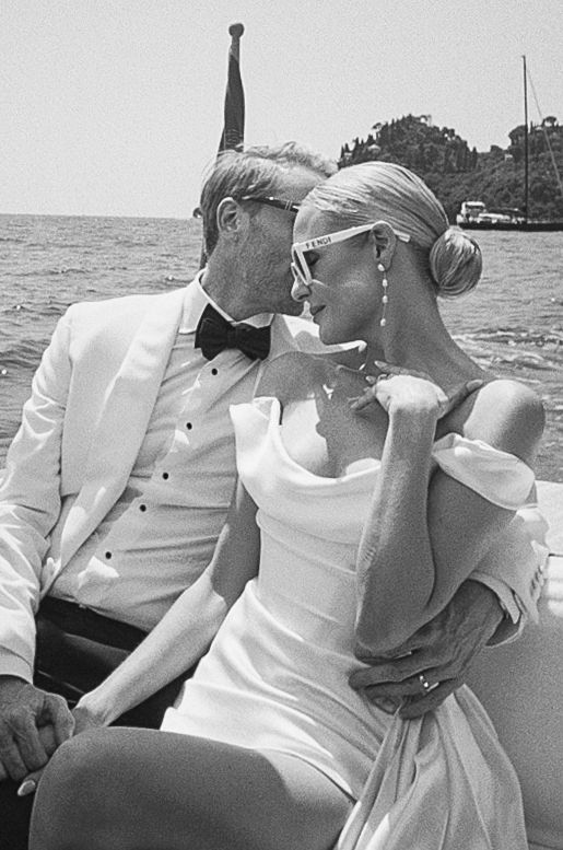 Black and white romantic portrait of newlyweds on boat, bride in off-shoulder gown with sunglasses.