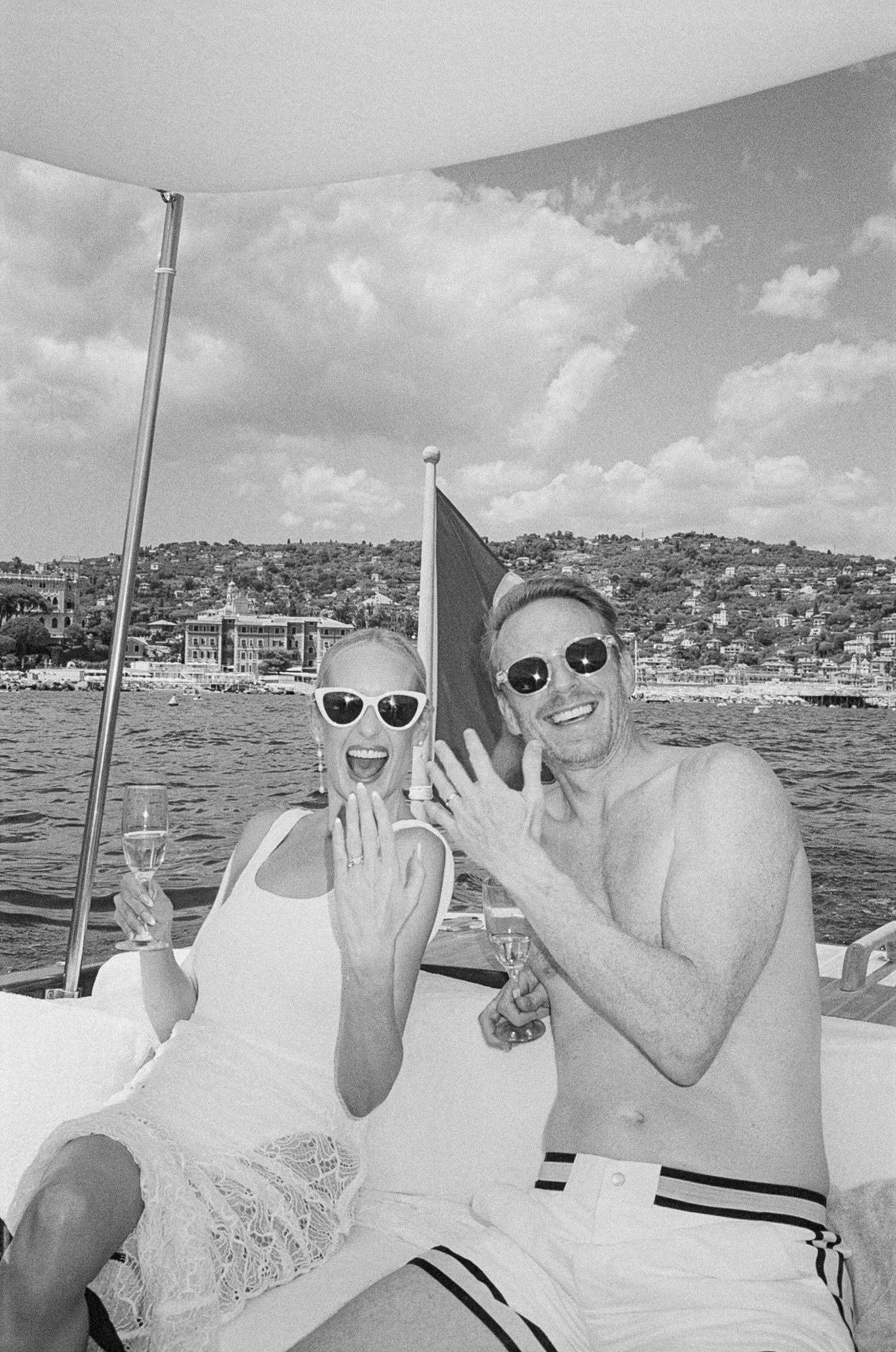 Joyful couple showing engagement rings on a boat with Italian coastal village in background.