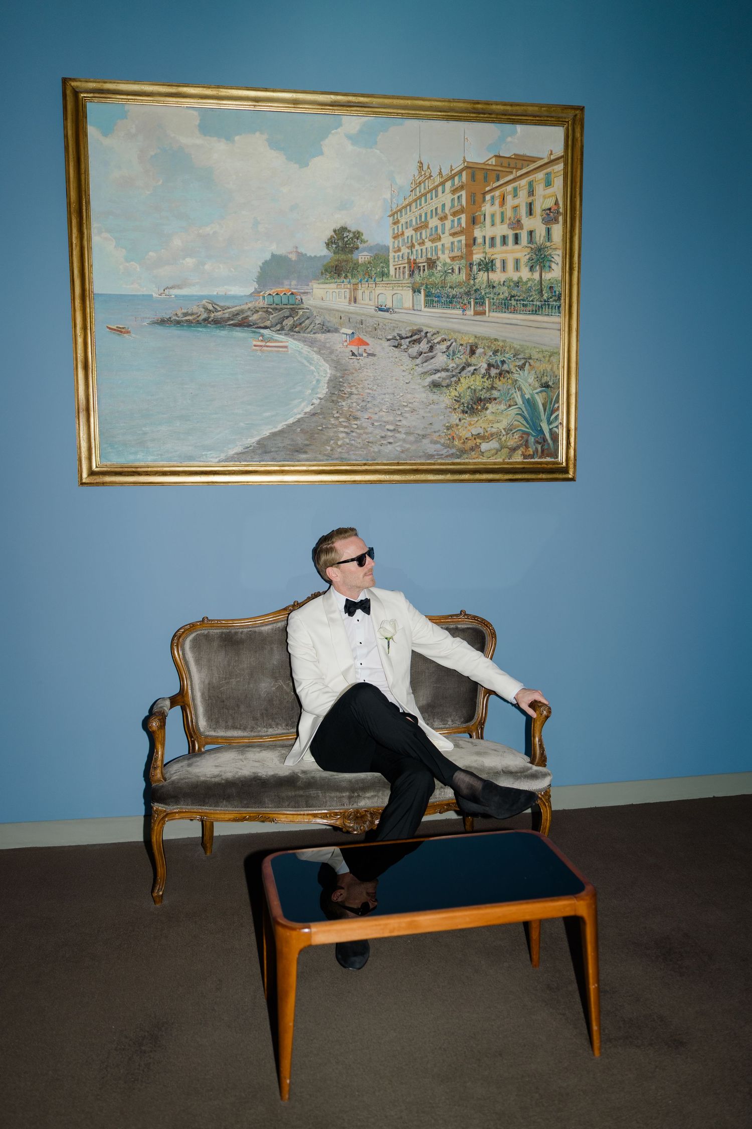 Groom in white tuxedo and sunglasses relaxing on vintage sofa beneath coastal landscape painting.