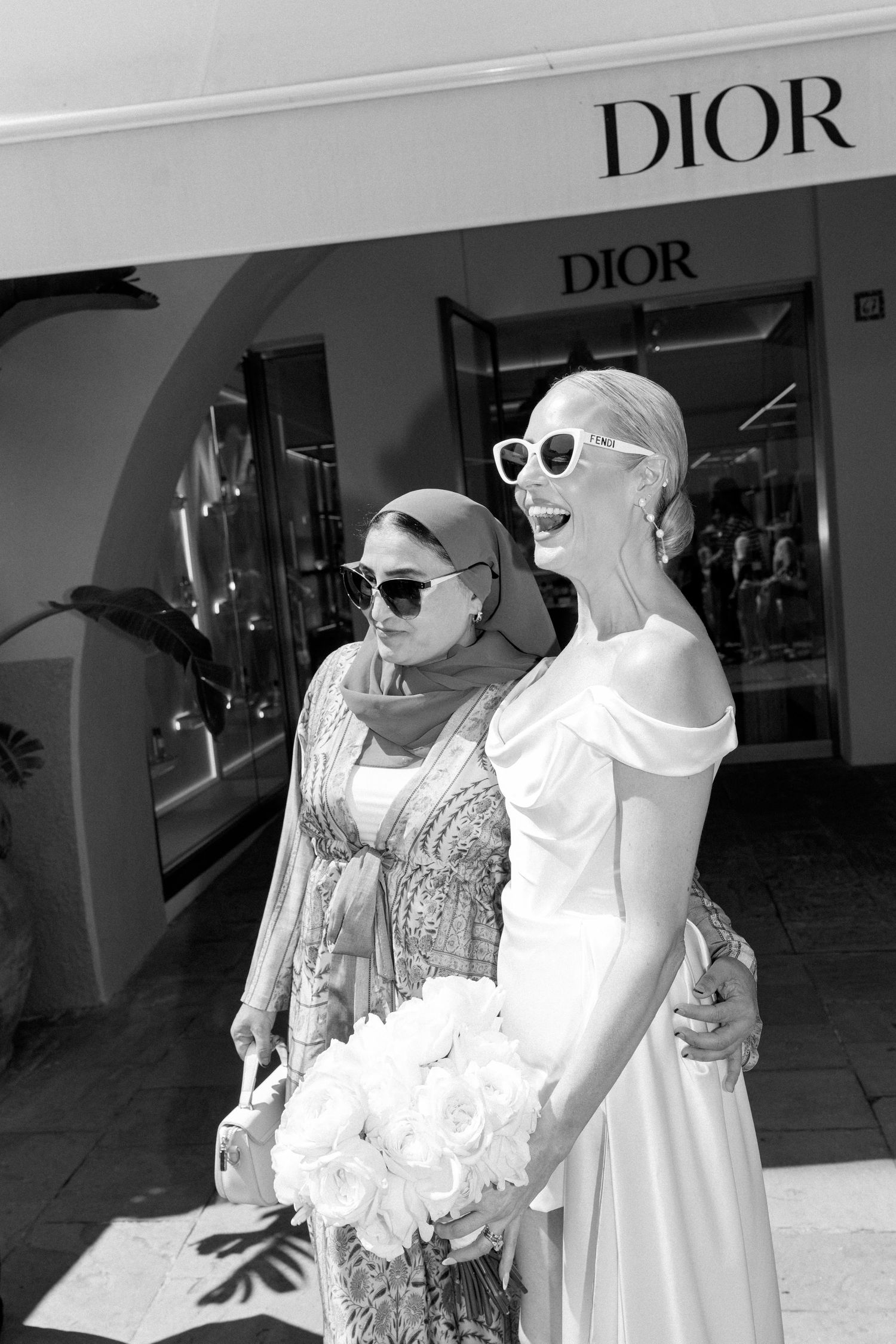 Bride in white gown holds bouquet near Dior store with guest in hijab, black and white photo.