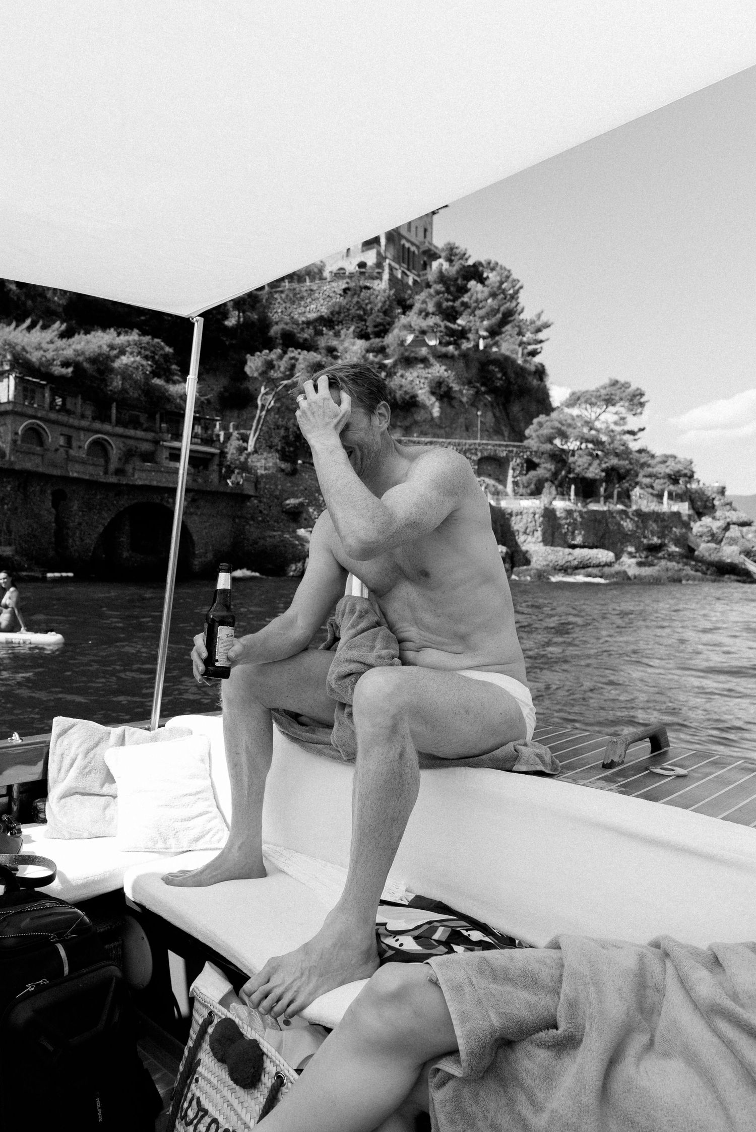 Black and white photo of a shirtless man sitting on a luxury boat near a rocky Mediterranean coastline.