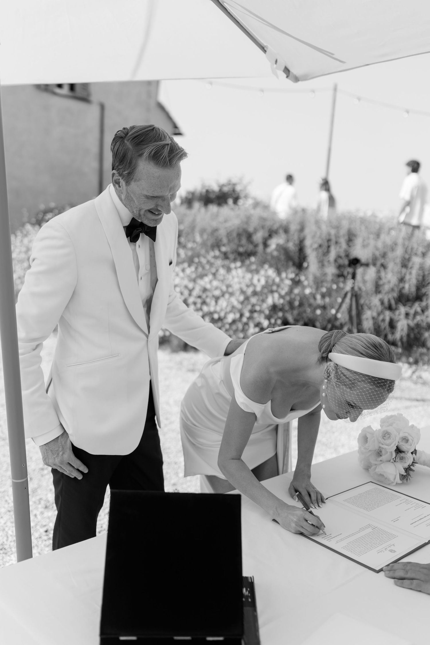 Bride signs wedding certificate as groom in white tuxedo watches during outdoor ceremony.