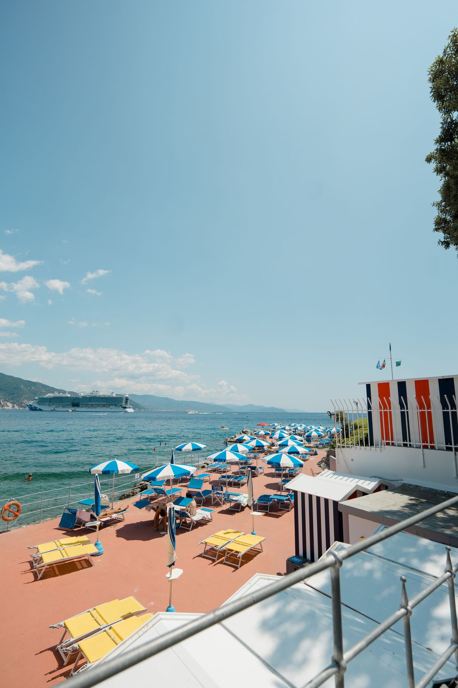 Sunny Italian Riviera beach with blue and white striped umbrellas, yellow loungers, and cruise ship in distance.