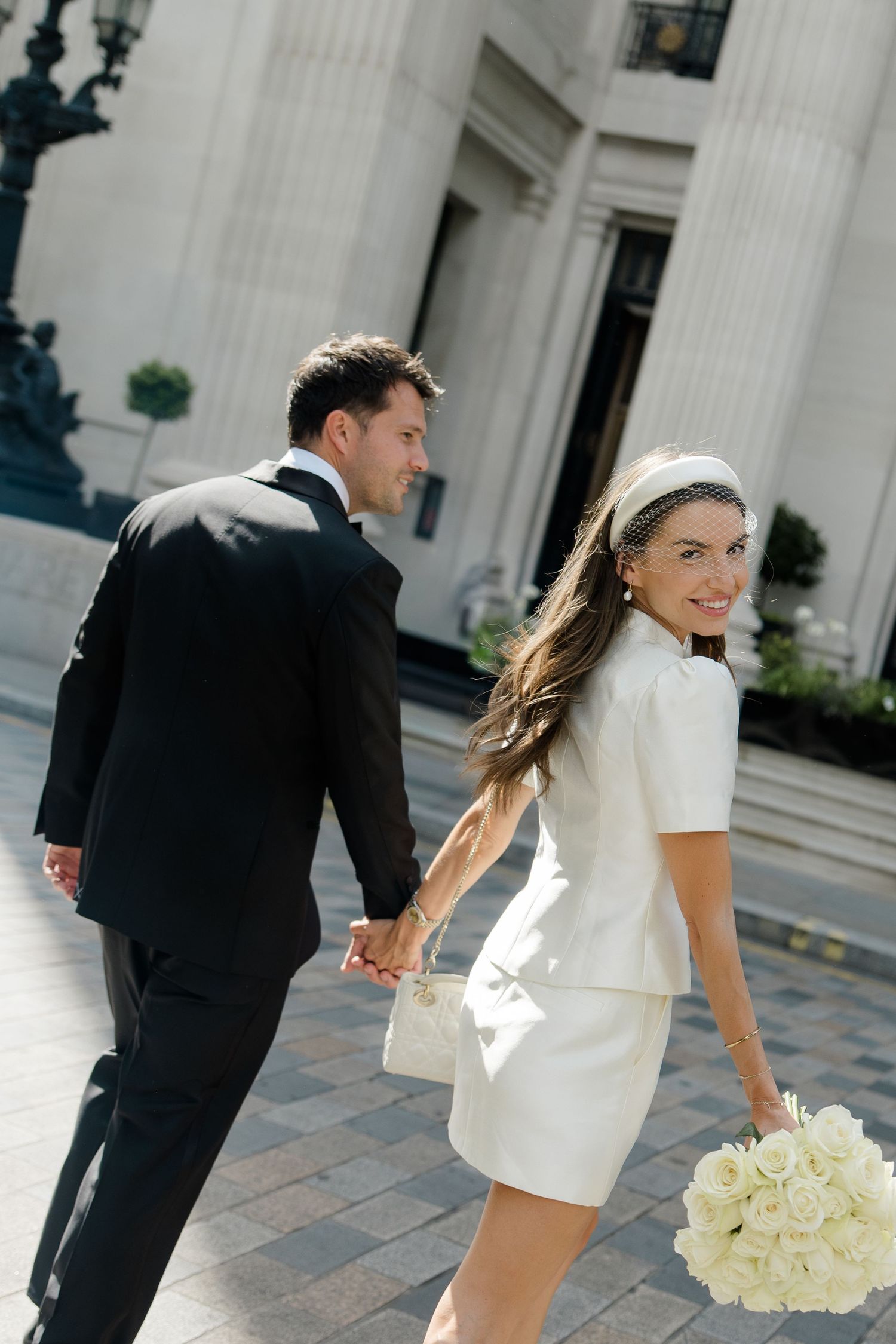 Smiling bride in white mini dress holds bouquet of white roses while walking hand-in-hand with groom in black suit.