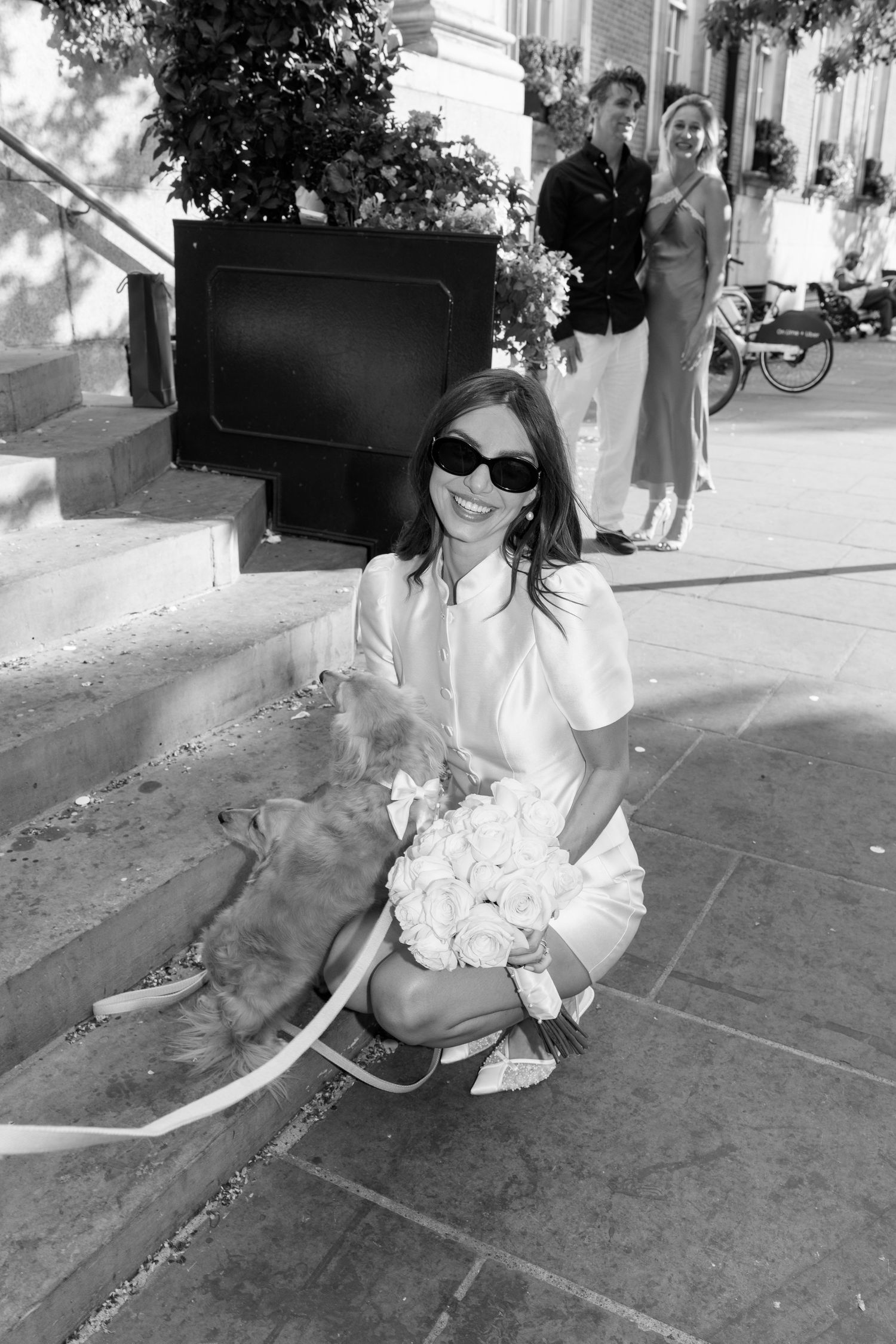 Smiling bride in sunglasses crouches with golden dog holding white bouquet on sunny steps.
