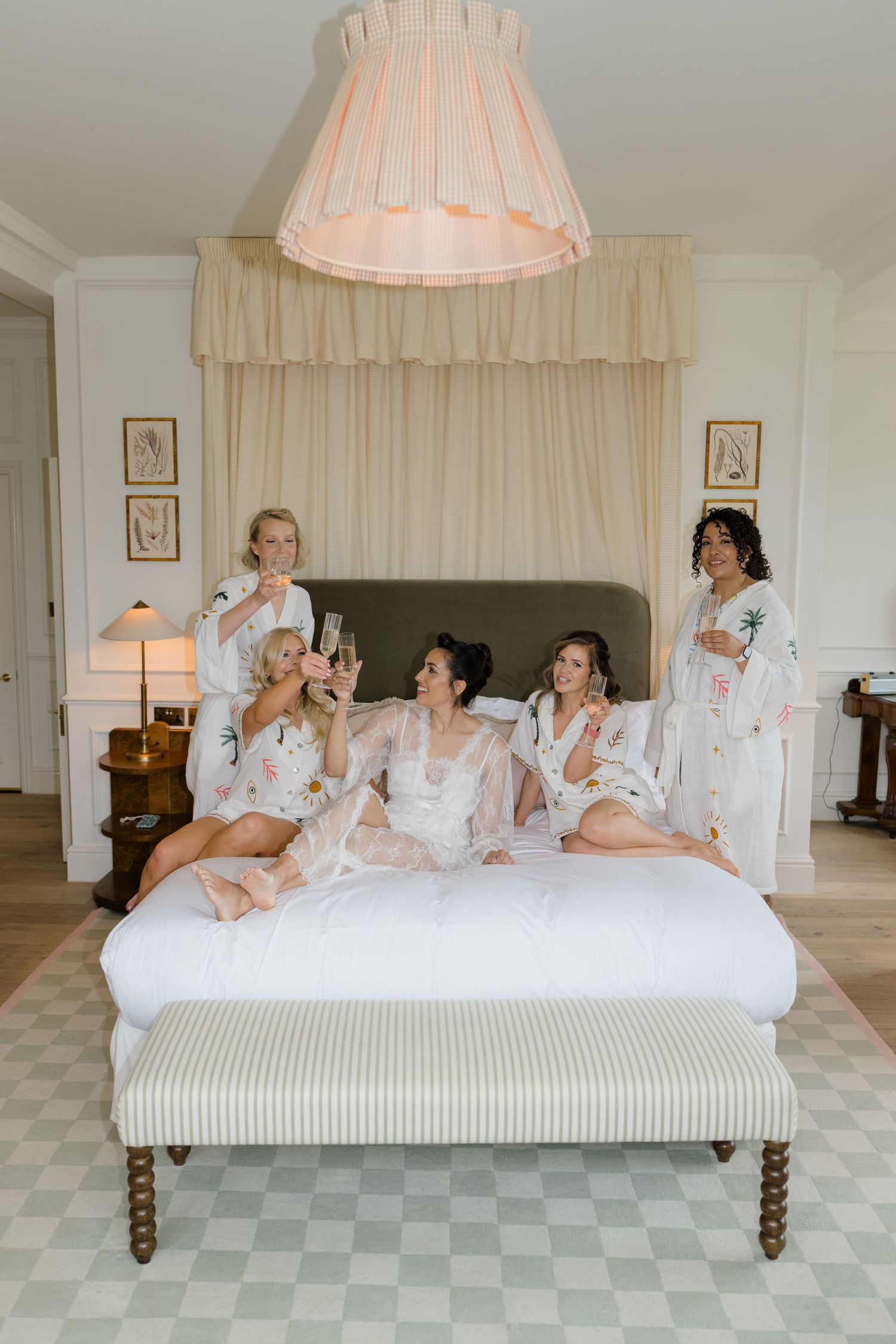 Bridesmaids in white robes celebrate with champagne on a luxurious bed in an elegant hotel suite.