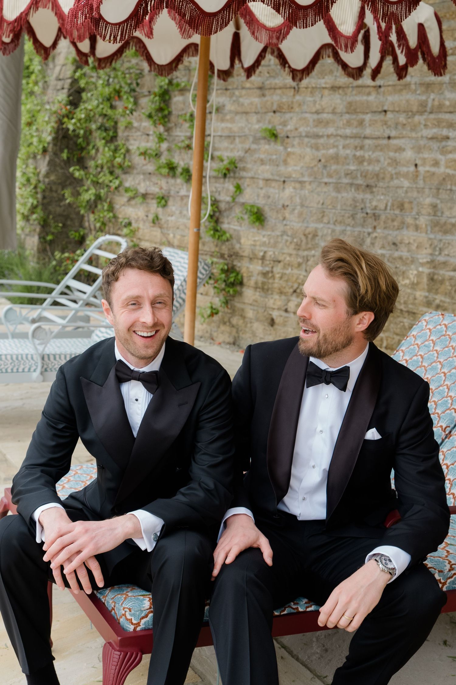 Two grooms in black tuxedos laughing together seated under a fringed umbrella outdoors.