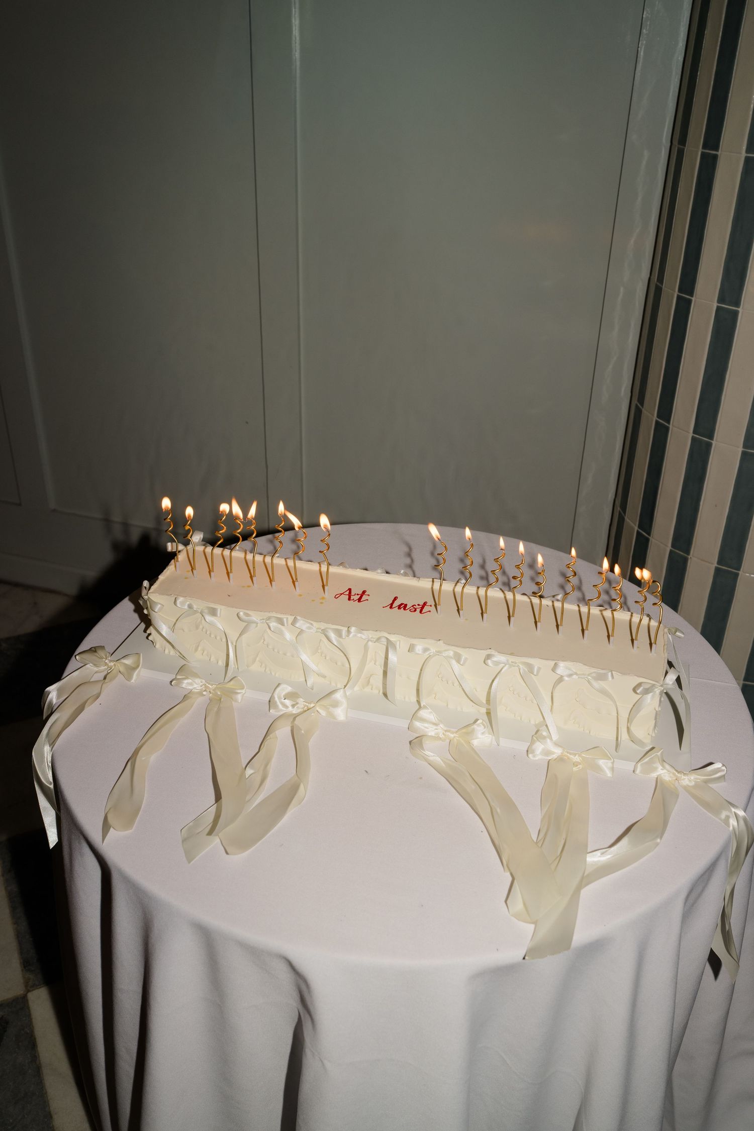 Elegant wedding cake with lit candles and white ribbon bows reading At Last on round table.