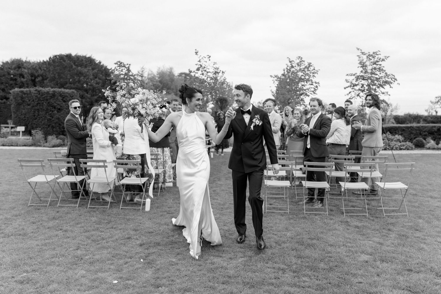 Black and white photo of newlyweds walking hand in hand past cheering guests after outdoor wedding ceremony.