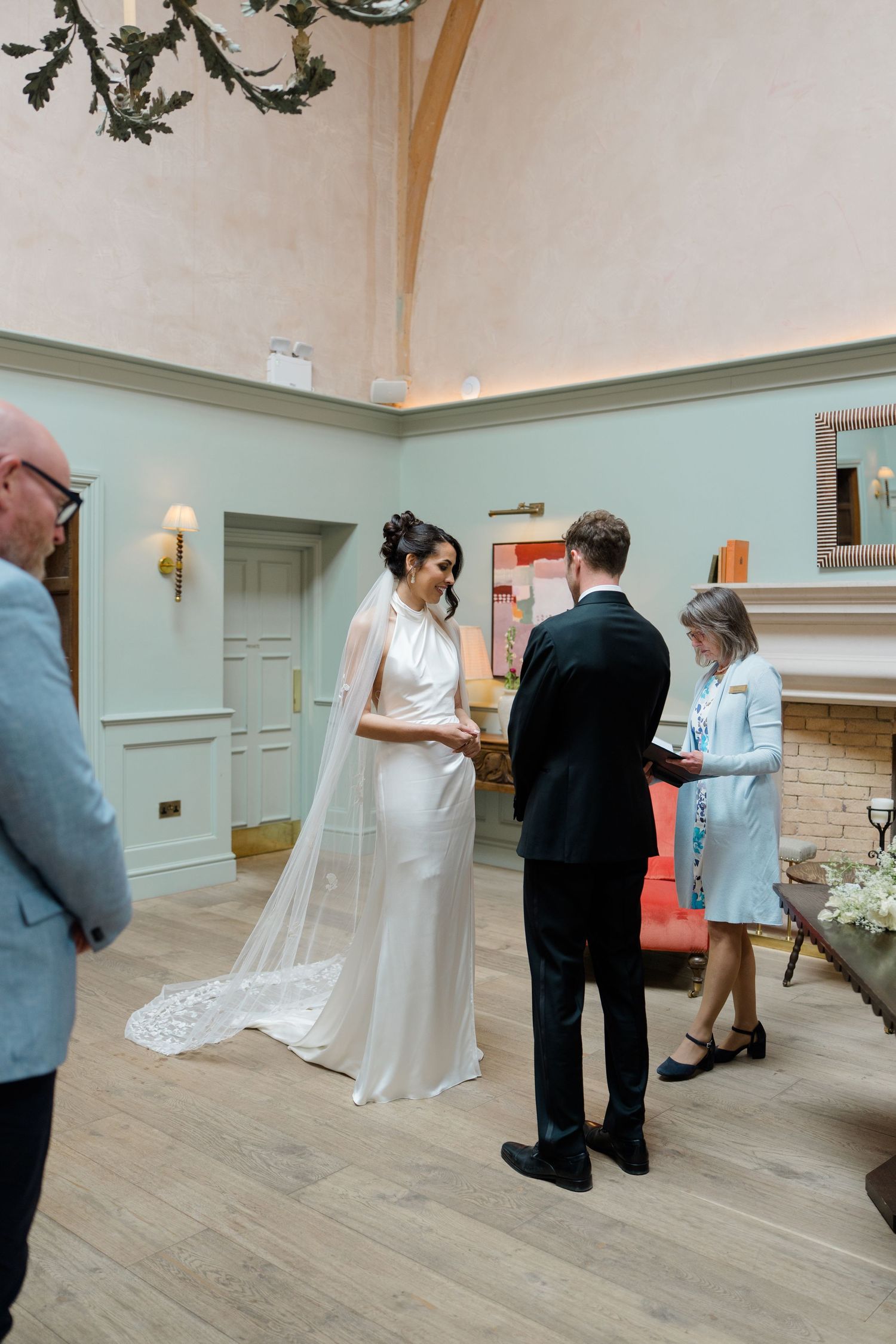 A bride and groom exchange vows indoors in an elegant room with high ceilings and a fireplace.