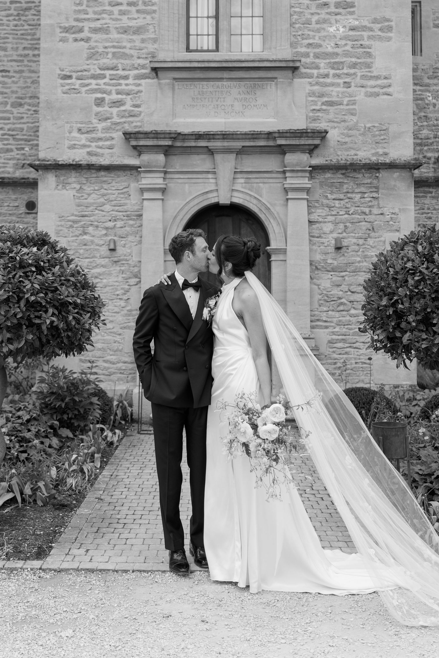 Bride and groom share a kiss outside a historic stone building, black and white wedding portrait.