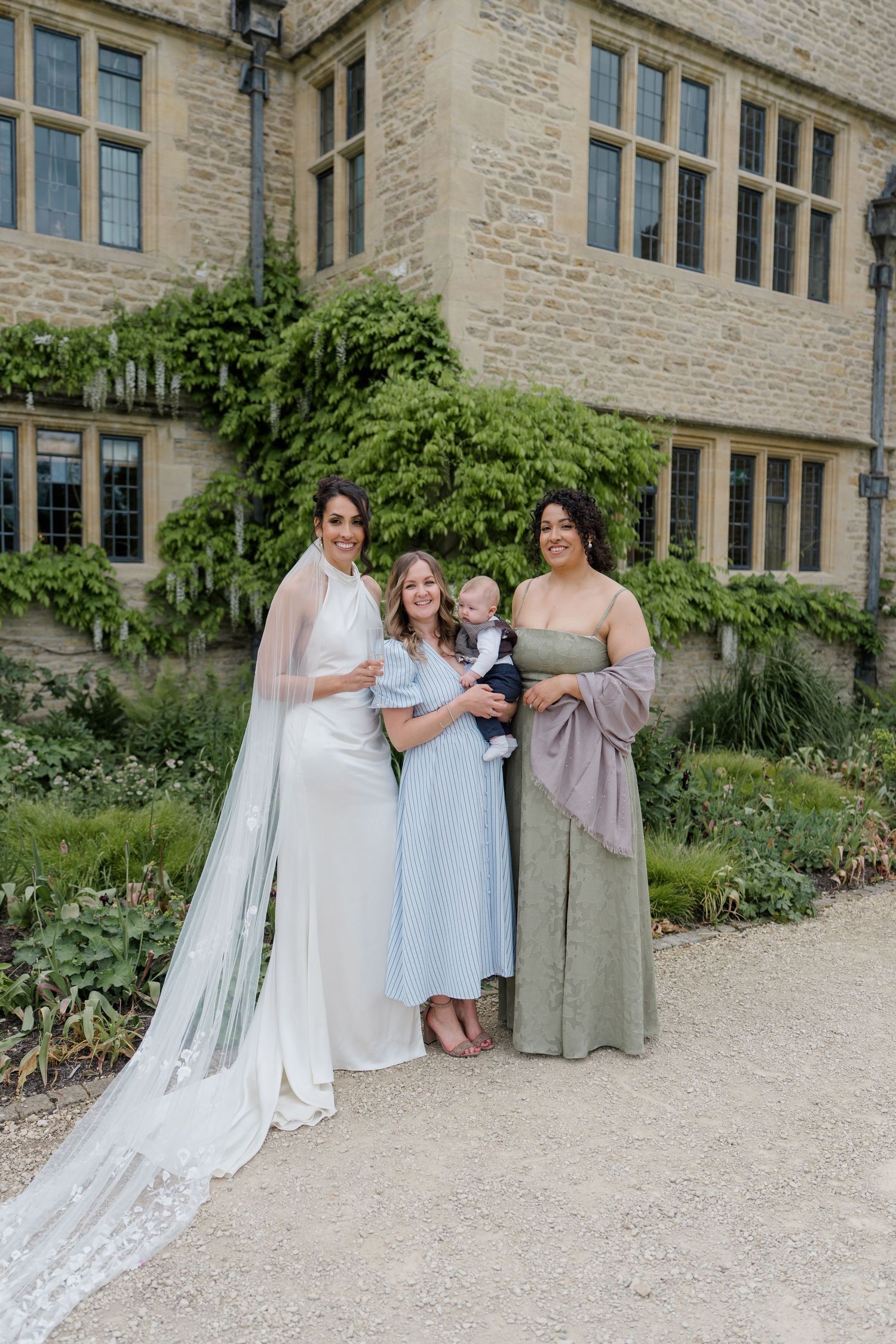 Bride in halter neck gown with long veil poses with two female guests and baby outside stone manor.