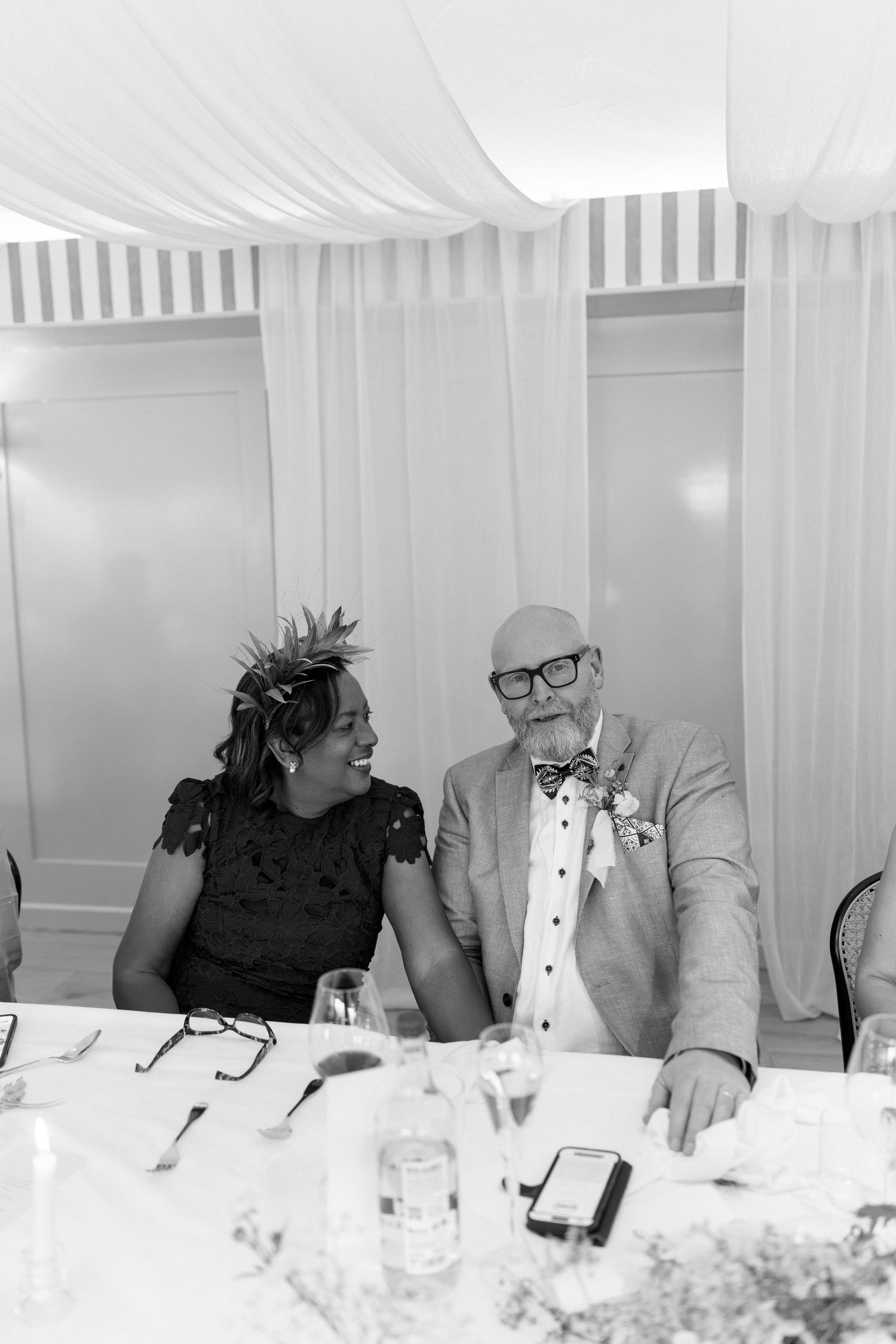 Black and white photo of smiling wedding guests, woman in floral headpiece and man in bow tie.