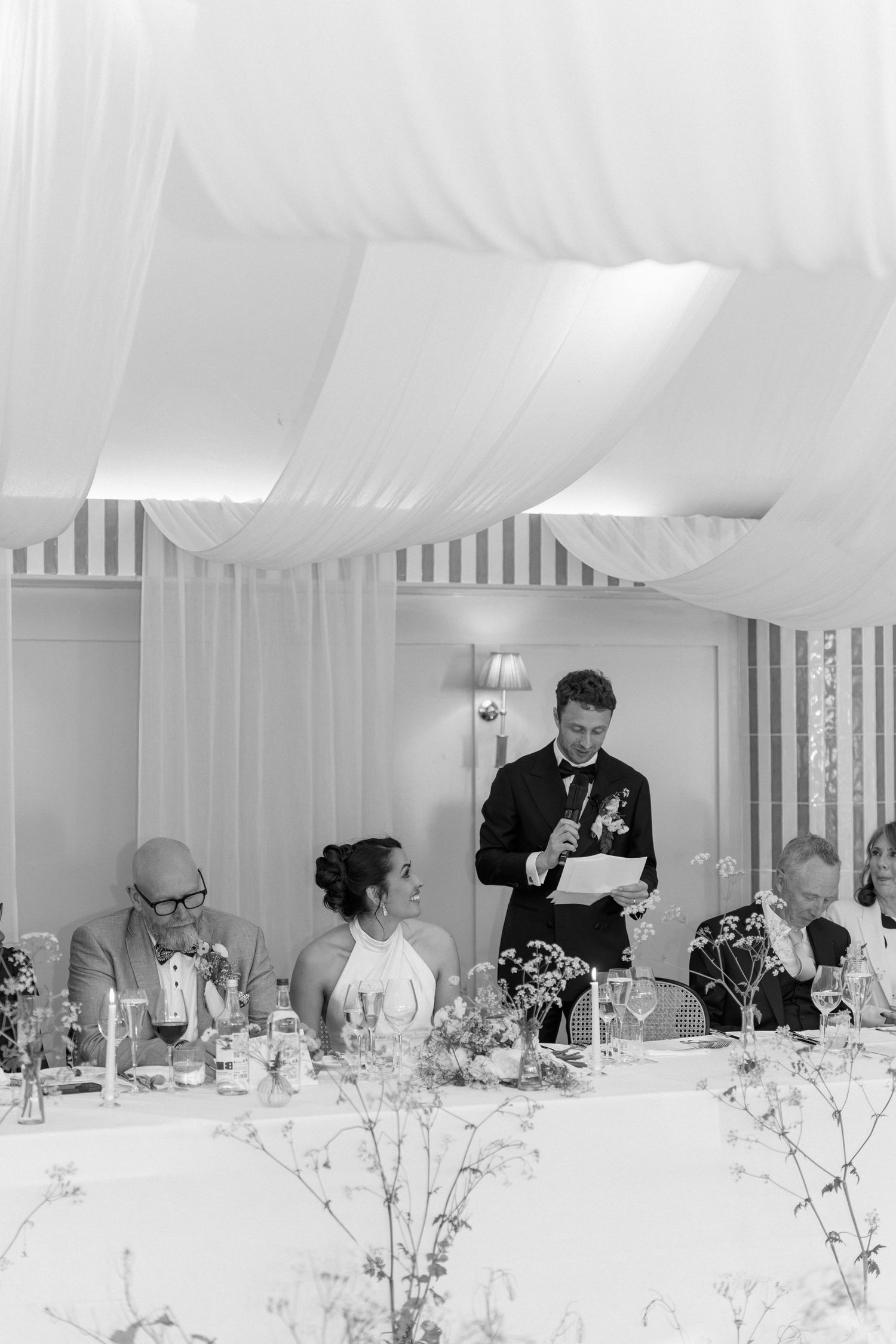 Black and white photo of a groom giving a speech at a wedding reception while guests smile and listen.
