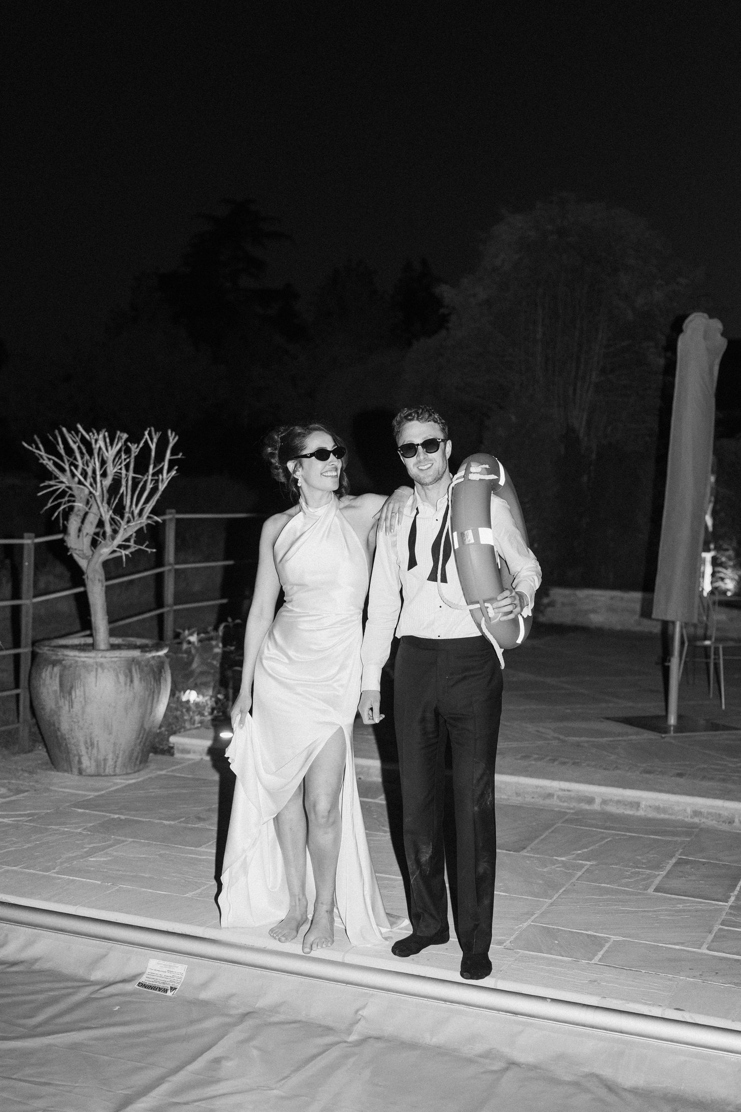 Black and white photo of a bride and groom in sunglasses posing by a pool at night.