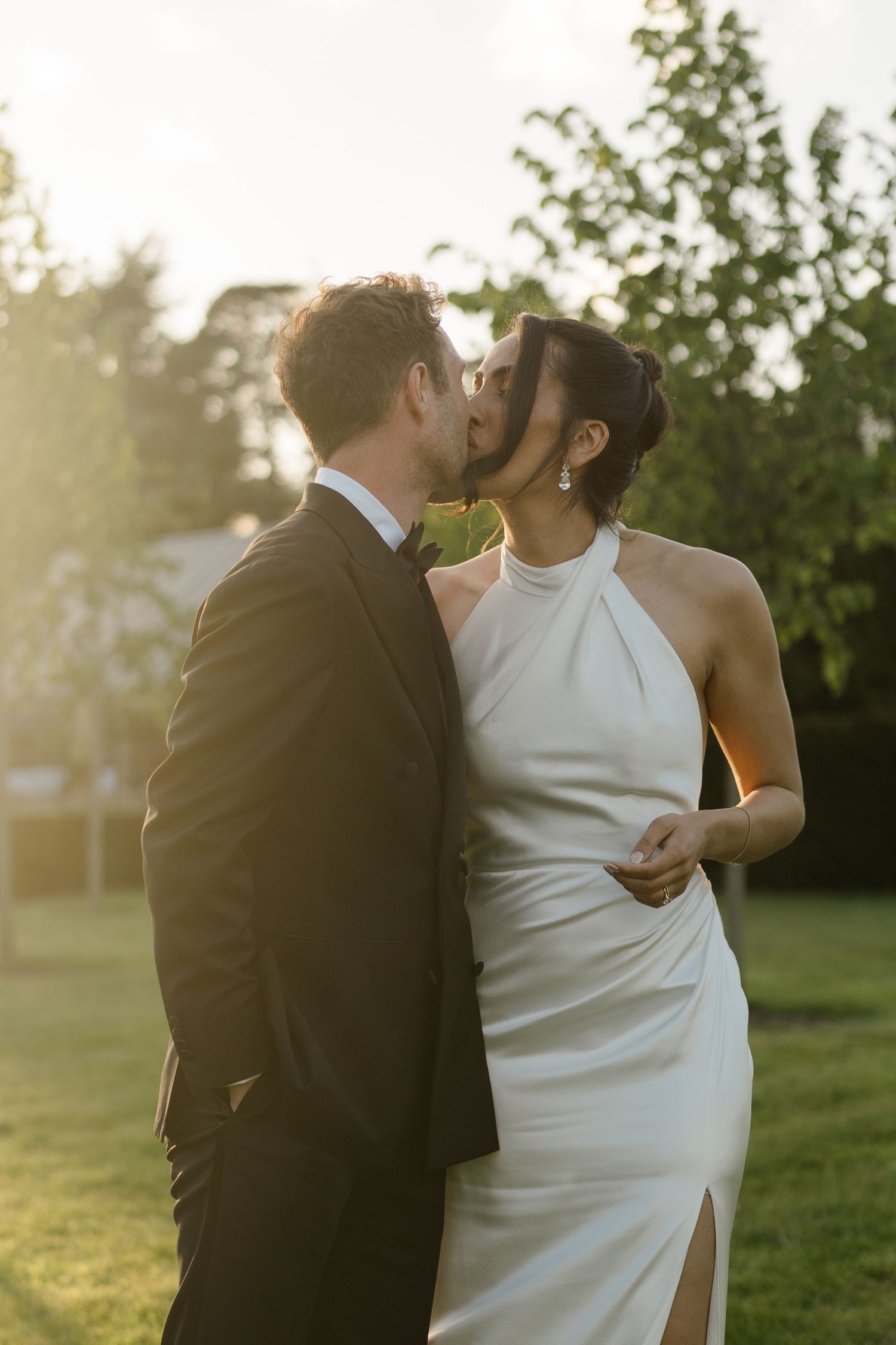 Bride and groom share a romantic golden hour kiss outdoors with soft sunlight filtering through trees.