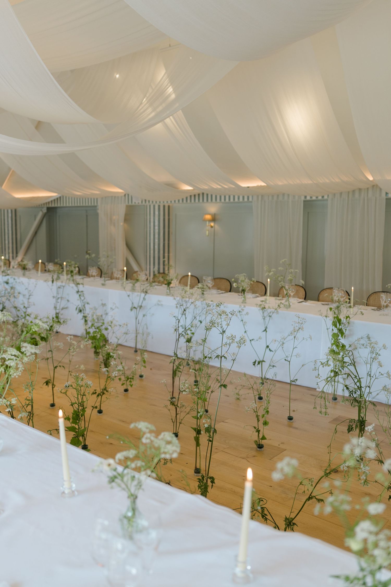 Elegant marquee wedding reception with long tables, white draping, candles, and delicate floral stems.