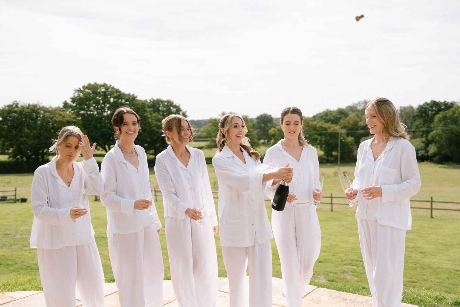A group in all-white outfits stands together on a sunny lawn celebrating with drinks.
