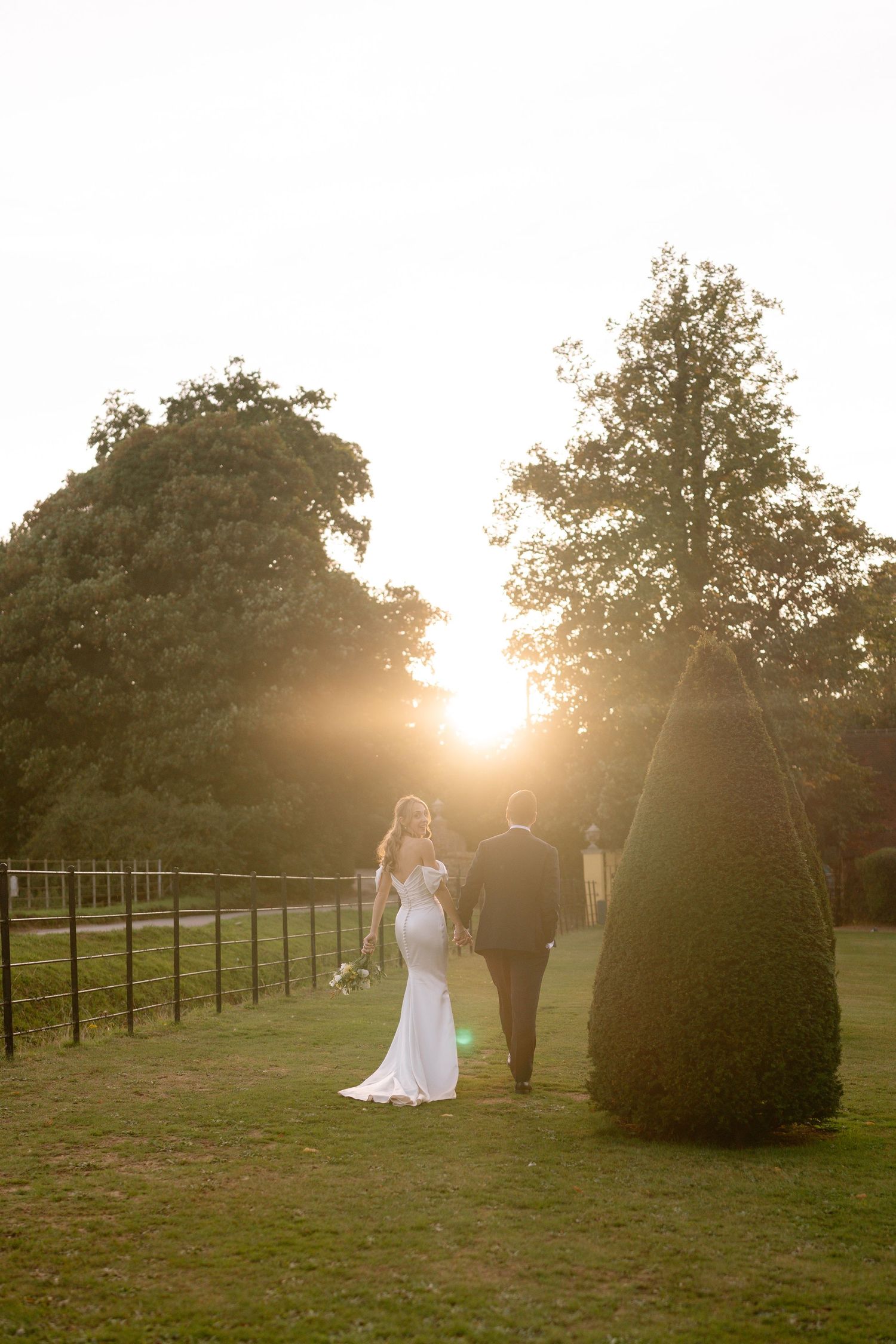 A wedding couple walks into the sunset through a manicured garden with topiary trees.