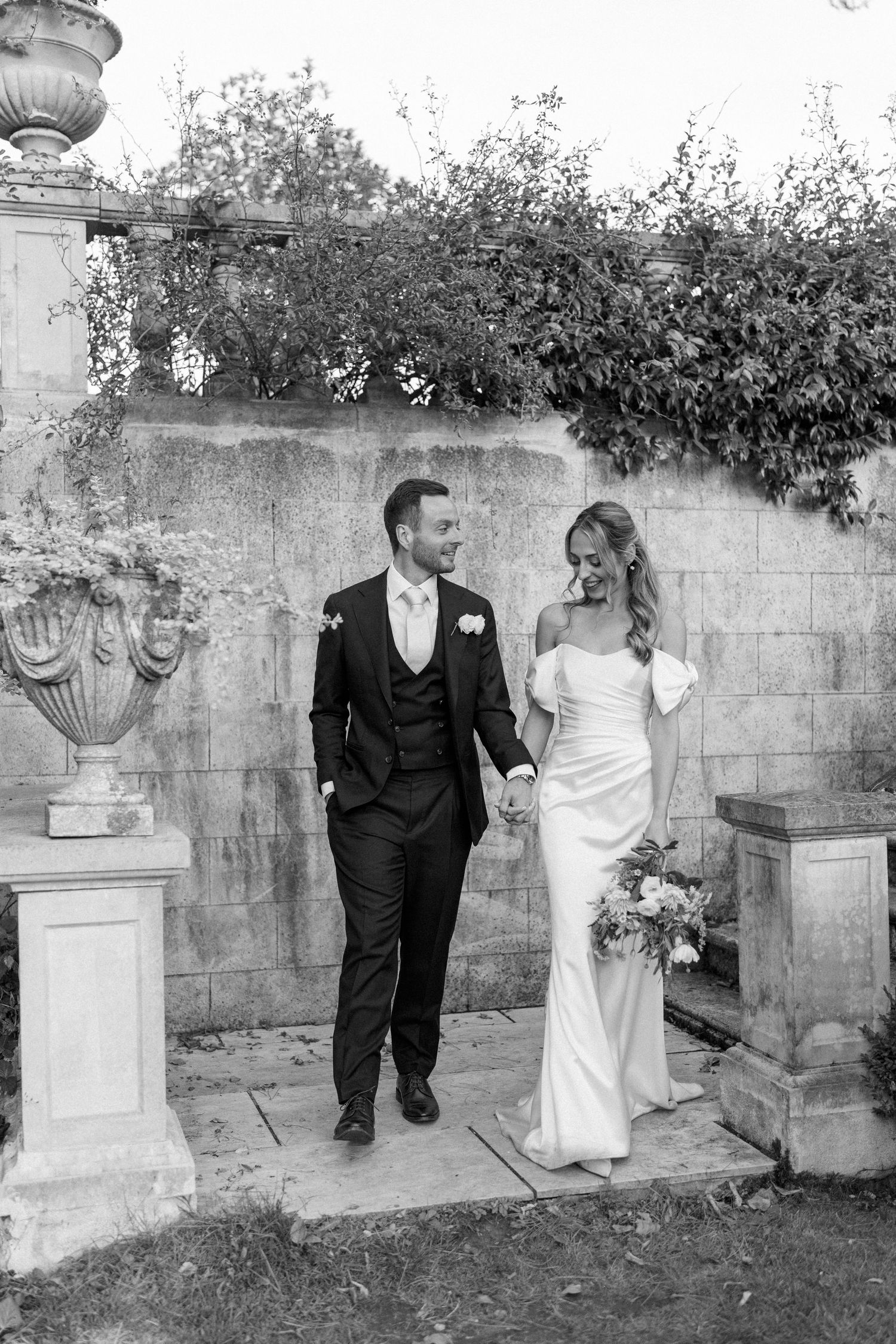 Couple walking along stone wall with decorative urns and garden landscaping.