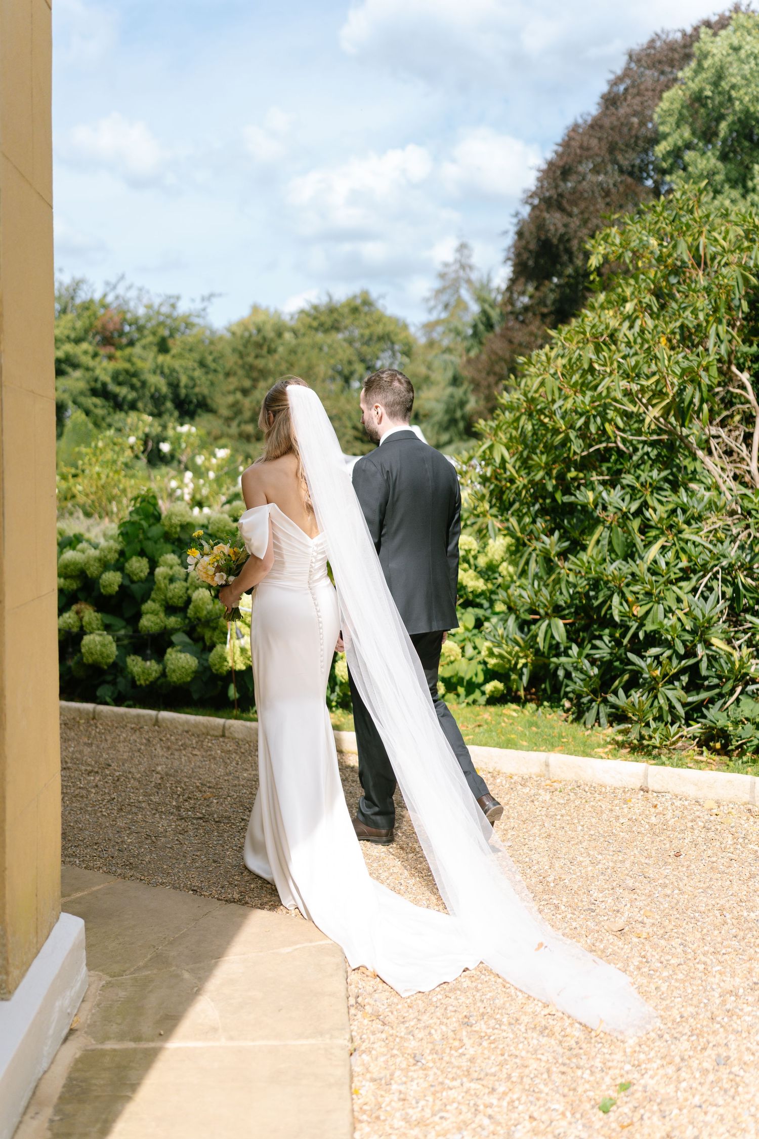 Wedding couple strolls through garden path near yellow building on sunny day.