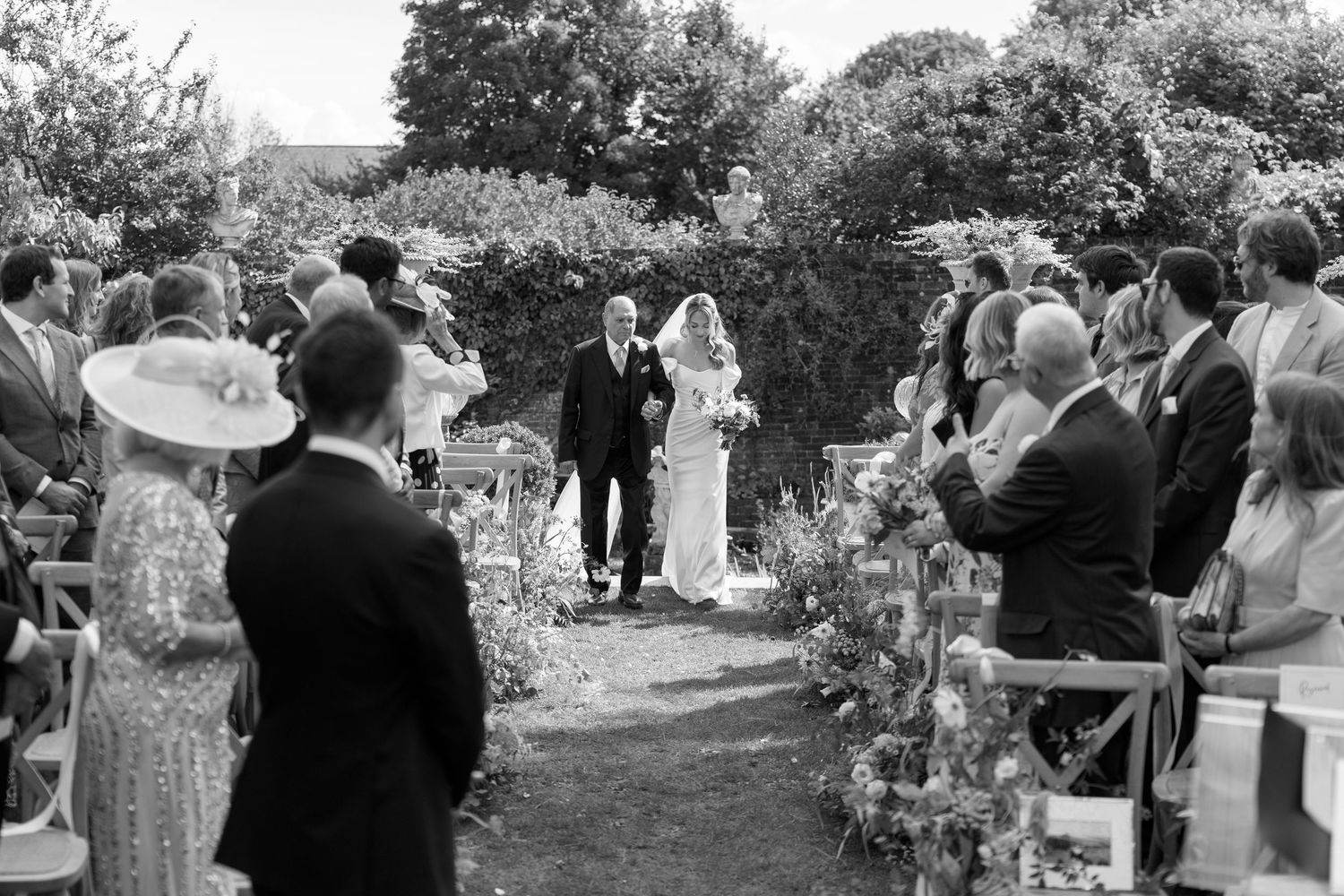 Wedding ceremony guests seated in rows of wooden chairs at an outdoor garden venue.
