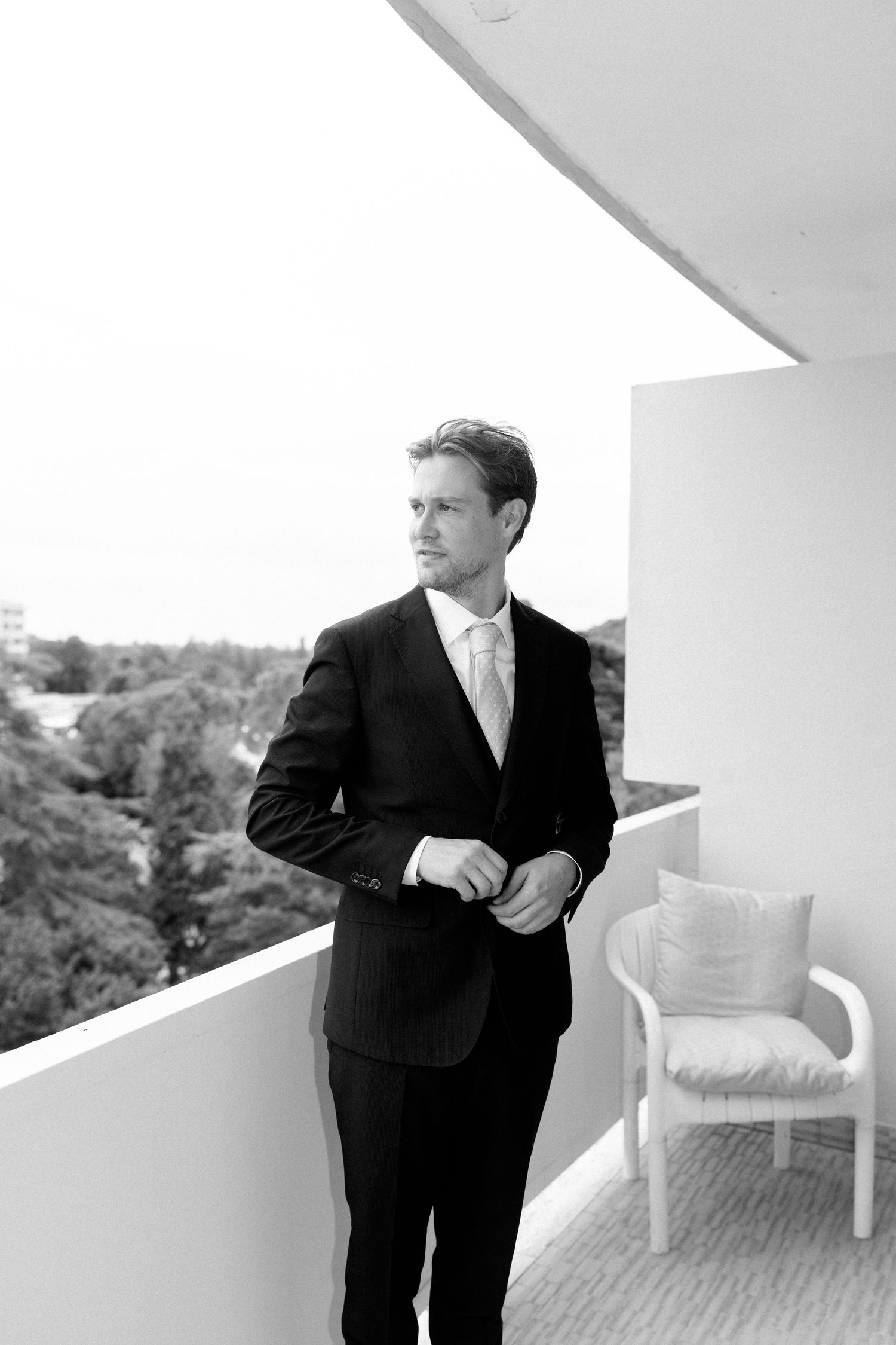 A person in formal attire stands on a balcony with white furniture and outdoor decor.