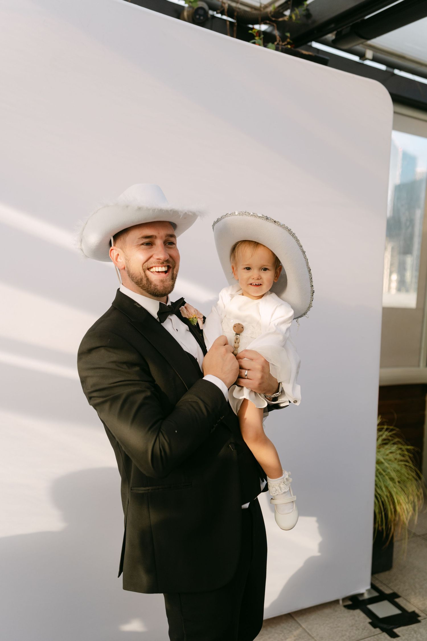 Wedding guest in formal attire holding small child in white outfit against white wall.