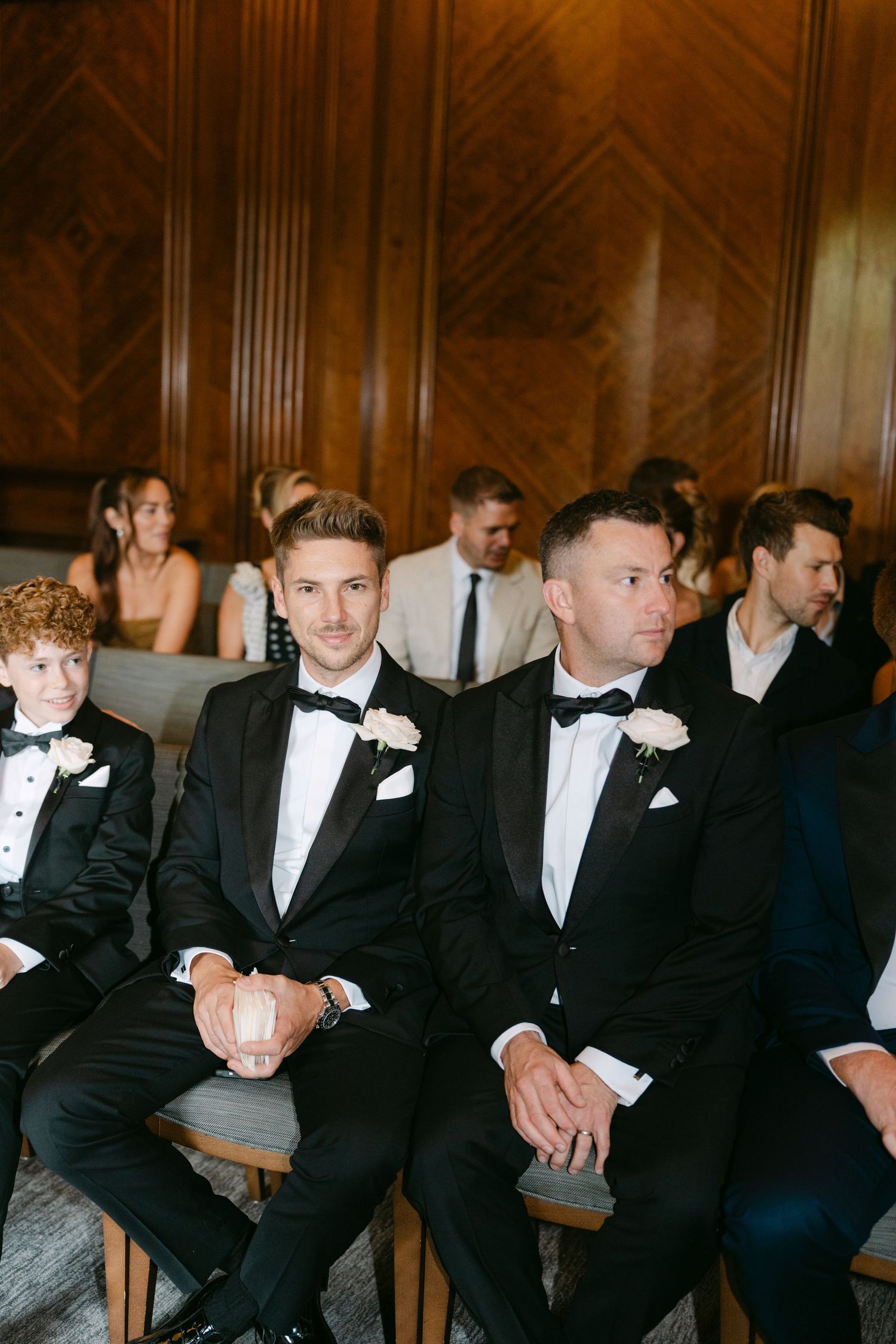 Groomsmen in black tuxedos sit in a row on wooden chairs in an elegant venue.