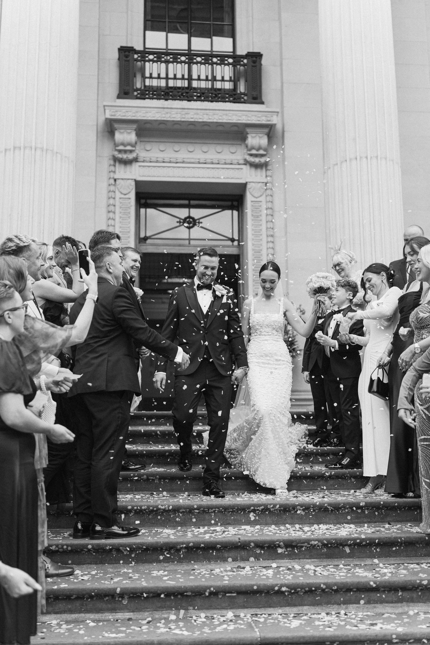 Wedding party celebrates with confetti toss on grand building steps.